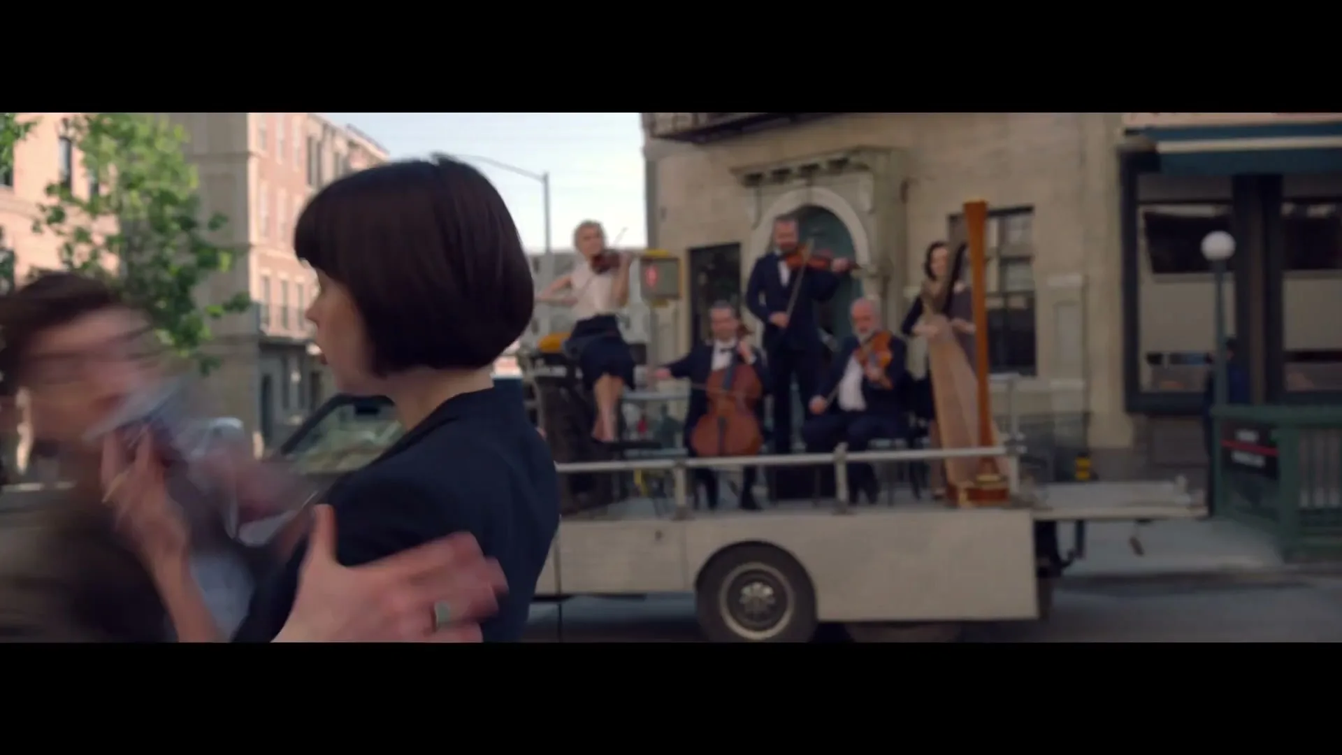 A man and woman embrace on a city street as an orchestra plays on the back of a truck, creating a romantic and cinematic moment.