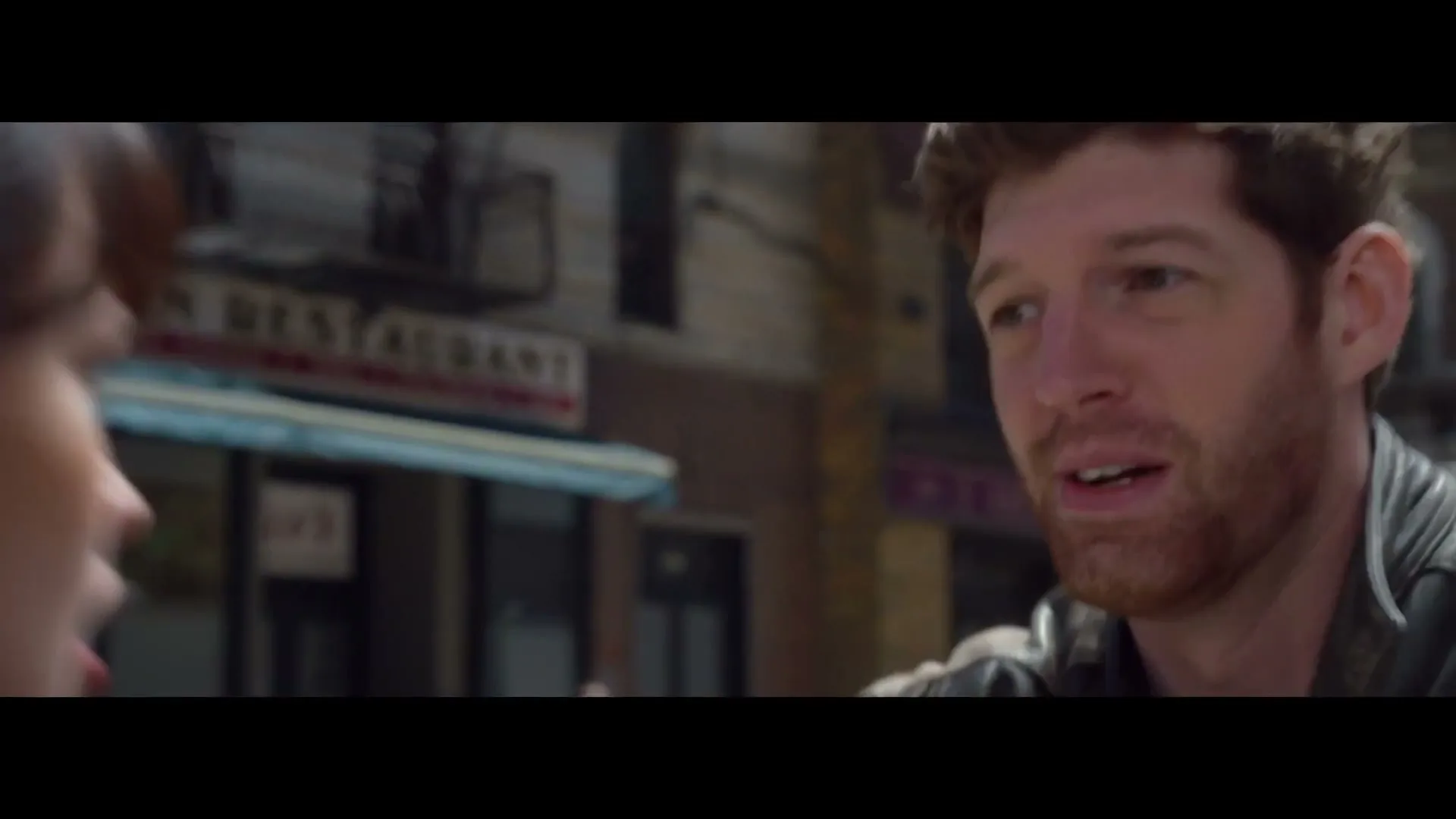 A man with a beard in a leather jacket talks to a woman, then runs away from a police car and other people on a city street.