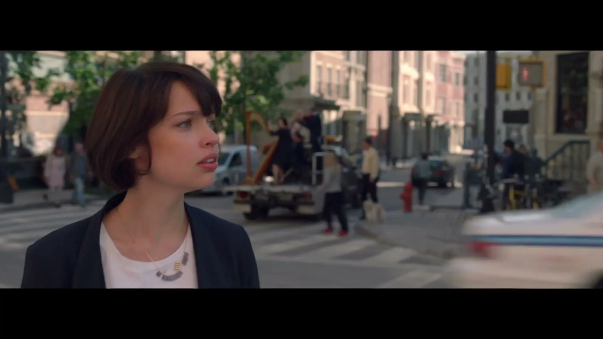 A young woman with short brown hair and a dark blazer looks surprised while standing on a city street during the day.