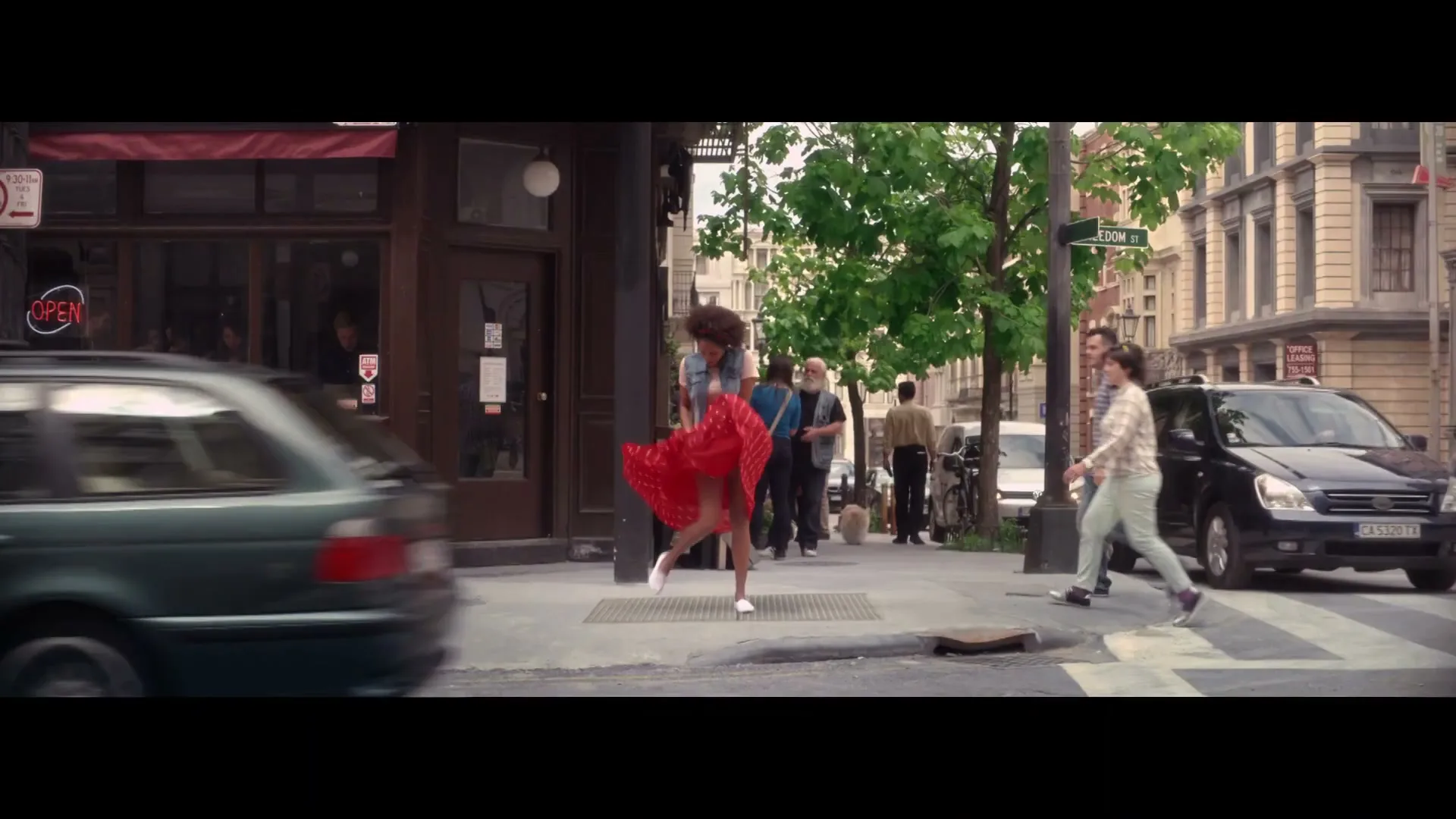 A young woman in a red polka dot dress stands on a city sidewalk, her dress blowing up in the wind, reminiscent of Marilyn Monroe.