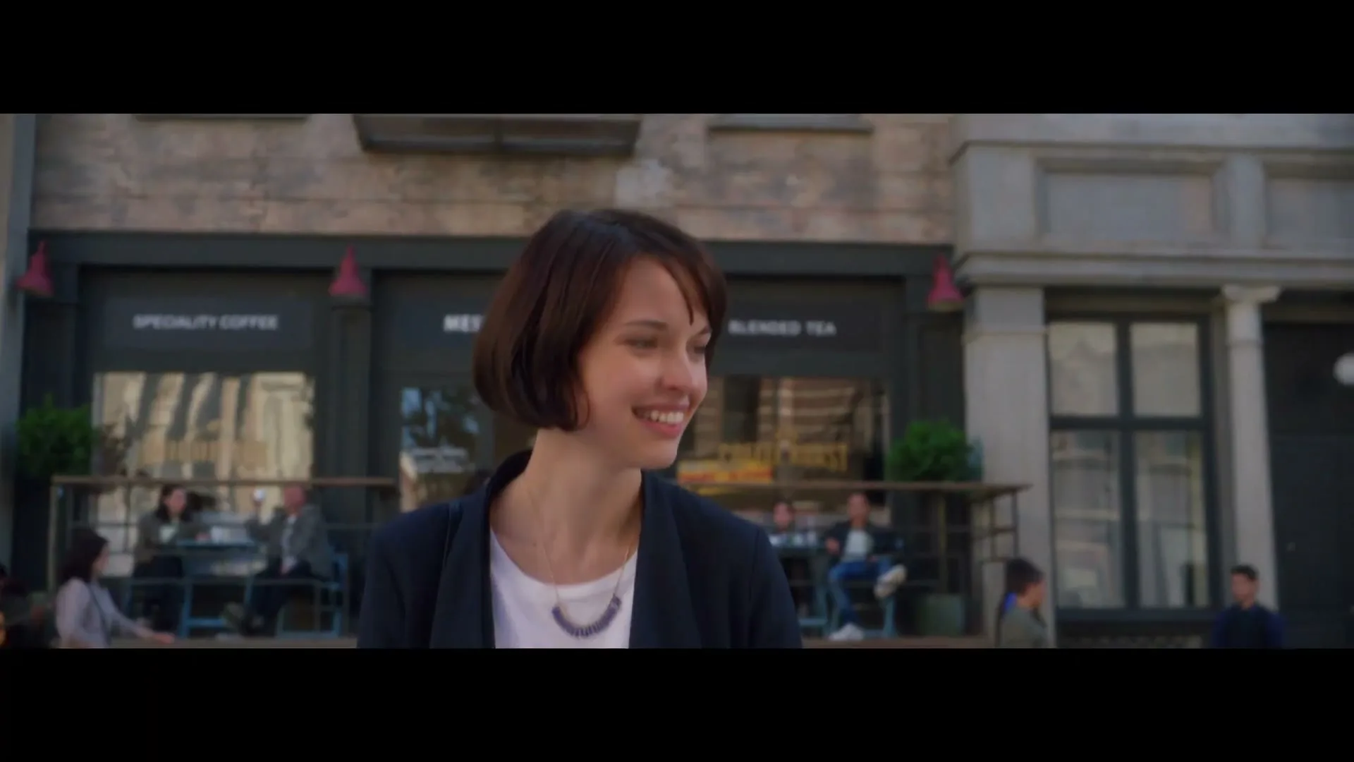 A young woman with short brown hair, wearing a dark blazer and white shirt, smiles while looking off-camera on a city street in front of a cafe.
