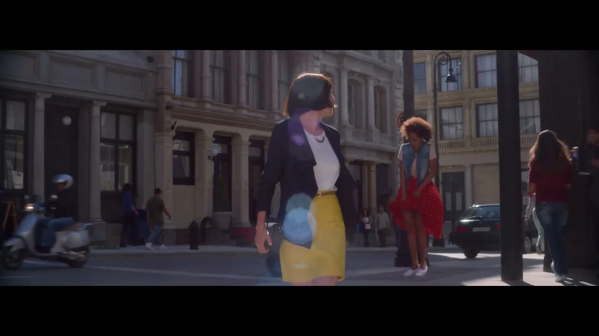 A woman in a yellow skirt and black jacket walks down a city street, smiling, as other people and vehicles pass by in the background.