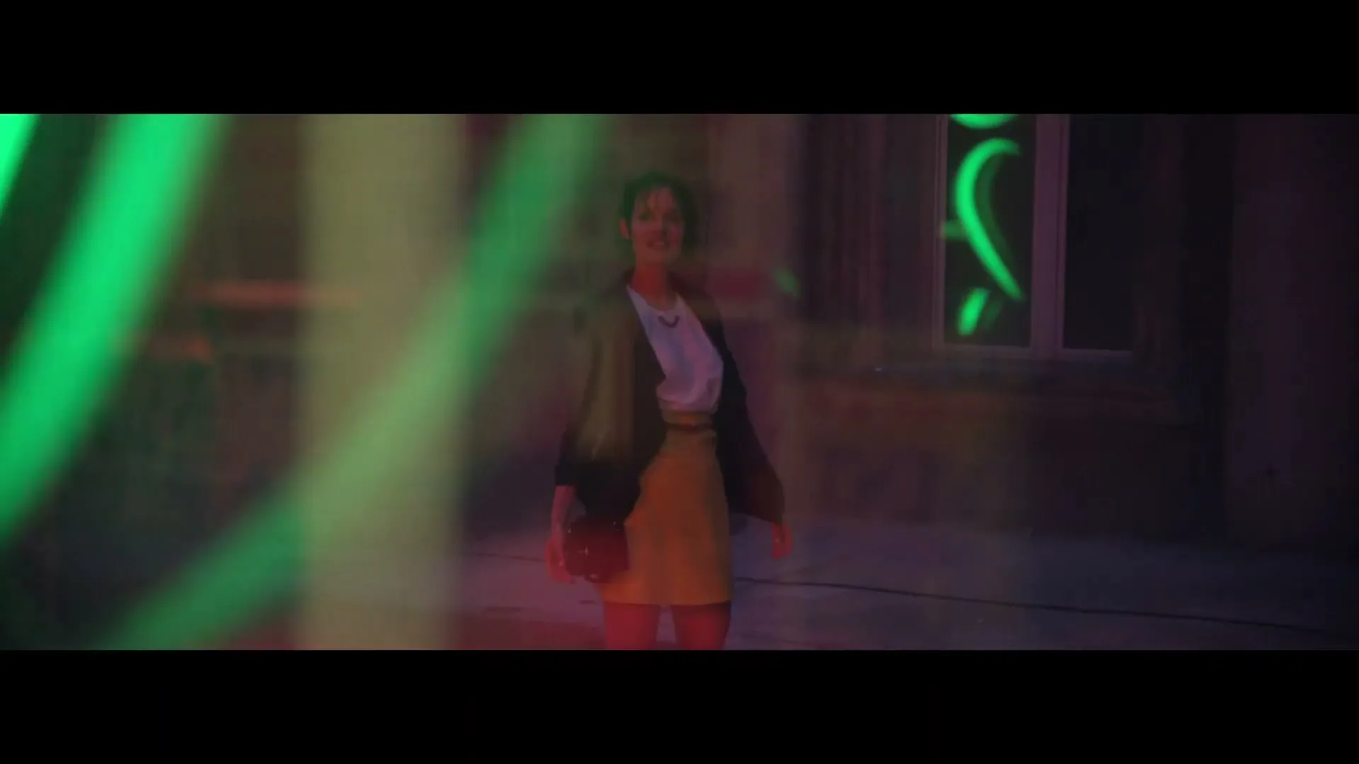 A young woman in a yellow skirt and dark jacket stands smiling in a dimly lit urban alleyway, with green neon lights reflecting in the foreground.