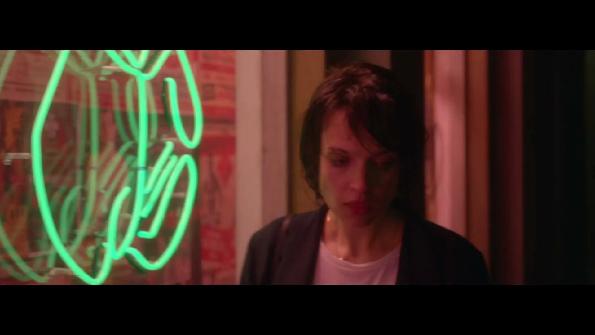A woman with short dark hair looks down, then up as someone applies makeup to her face, next to a green neon sign.