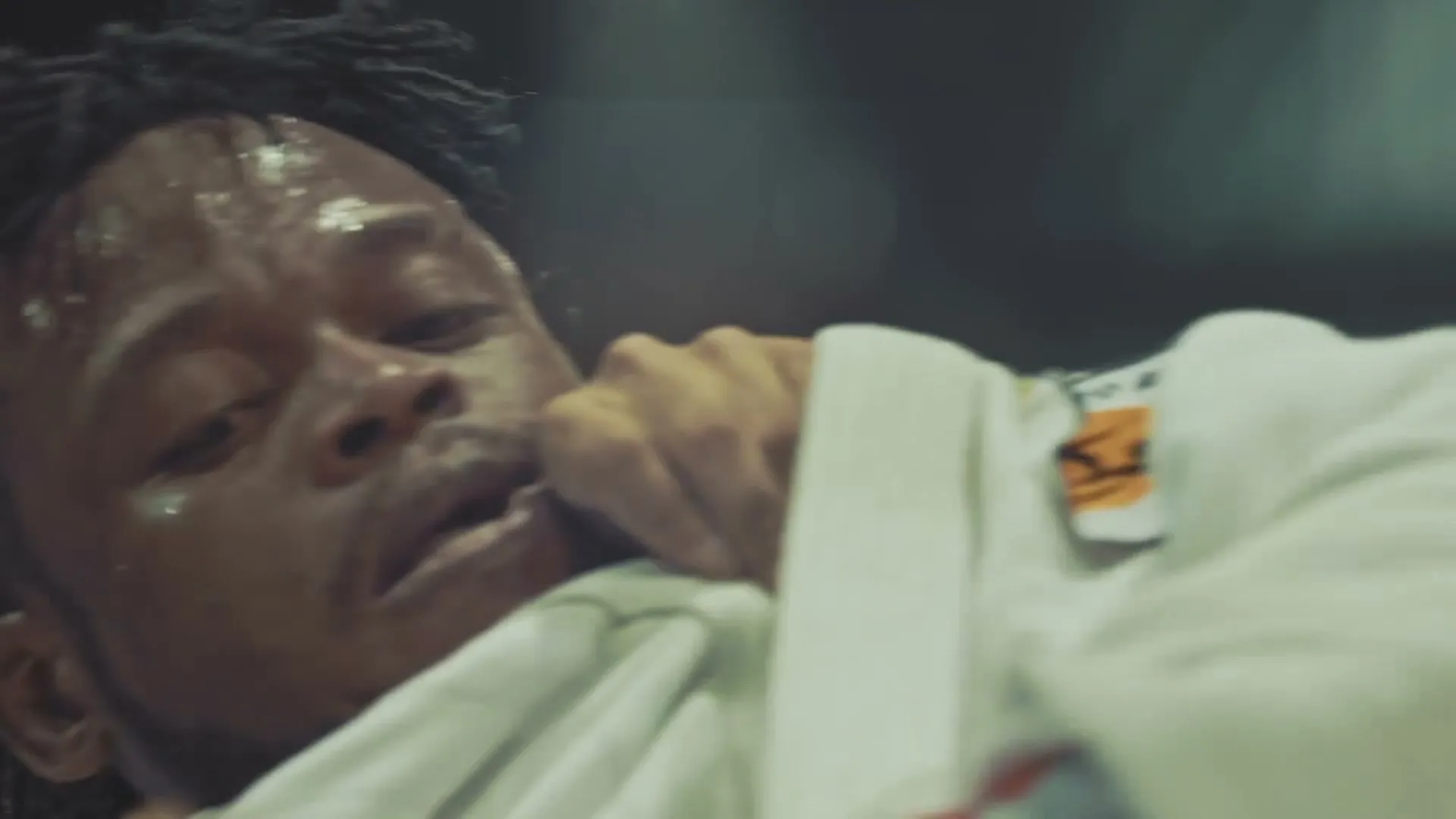 A young, sweaty male athlete with dreadlocks struggles in a white gi, grimacing in pain or exertion during a fight in a dimly lit arena.