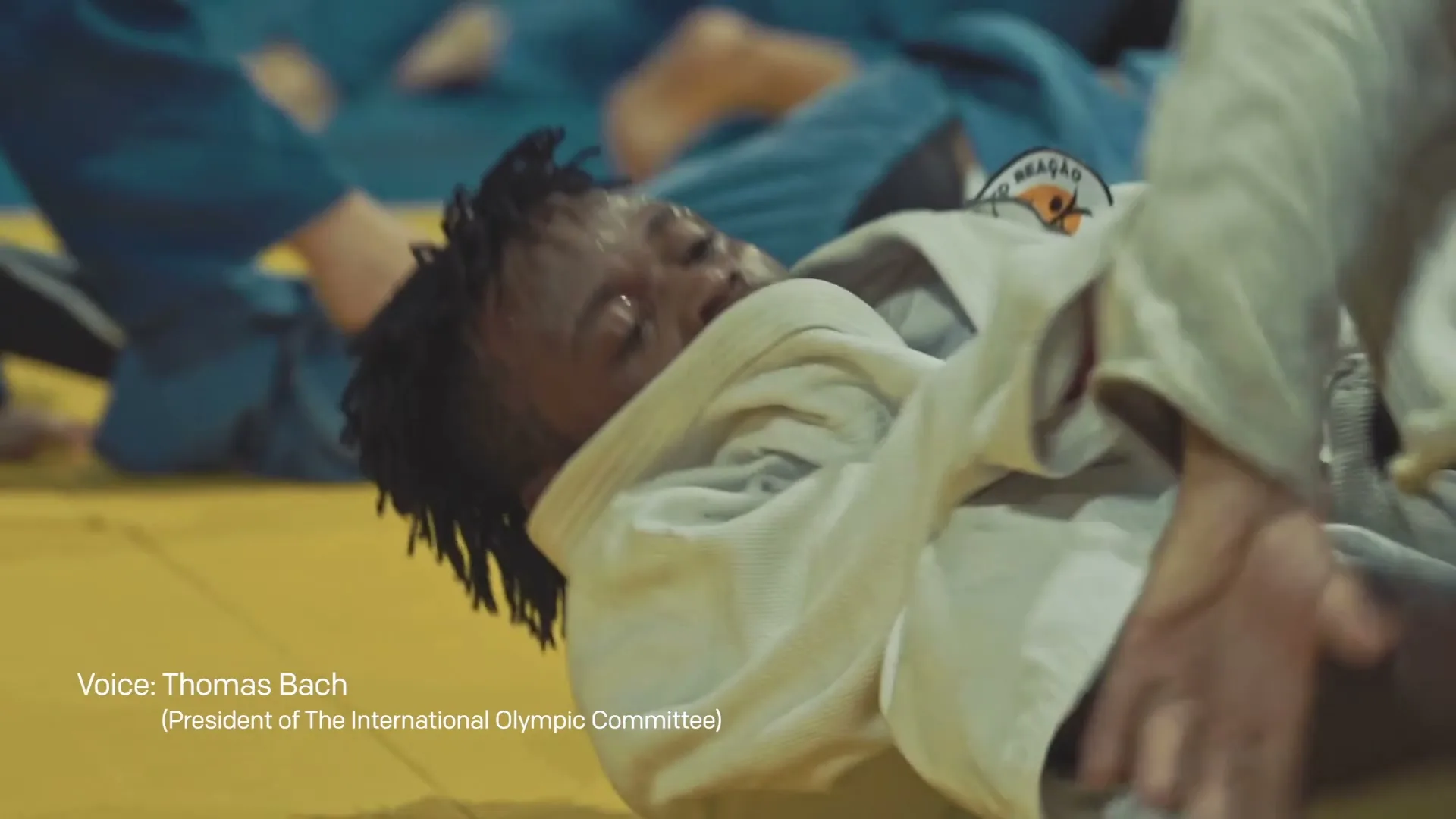 A male judoka with dreadlocks lies on a yellow mat, looking exhausted, while other judokas in blue uniforms are blurred in the background.