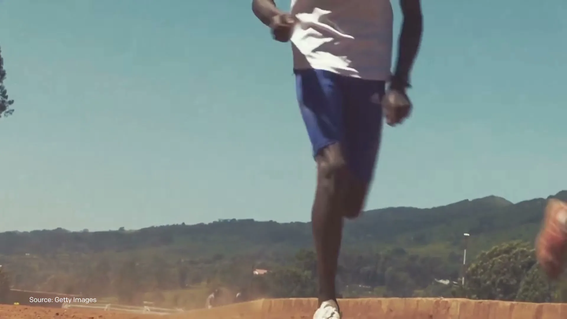 A group of male athletes in athletic wear run on a dirt track under a clear blue sky, kicking up dust as they move.
