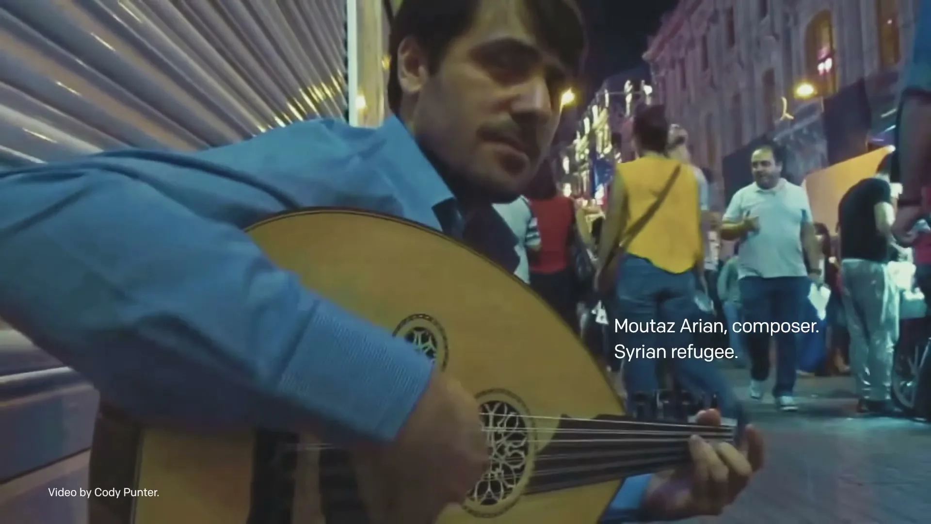 A male musician plays an oud on a busy city street at night, with a superimposition of sheet music appearing over the scene.