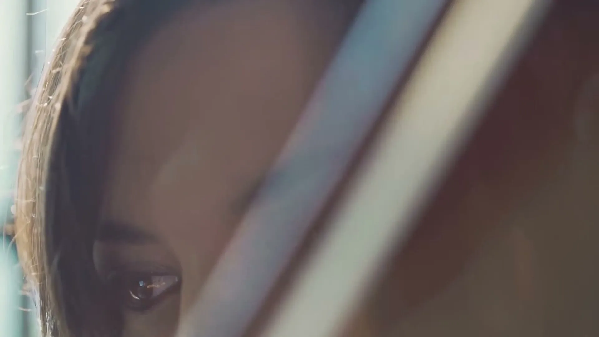 A woman with dark hair and eyes looks down, with a soft lens flare across her face, in an interior setting.