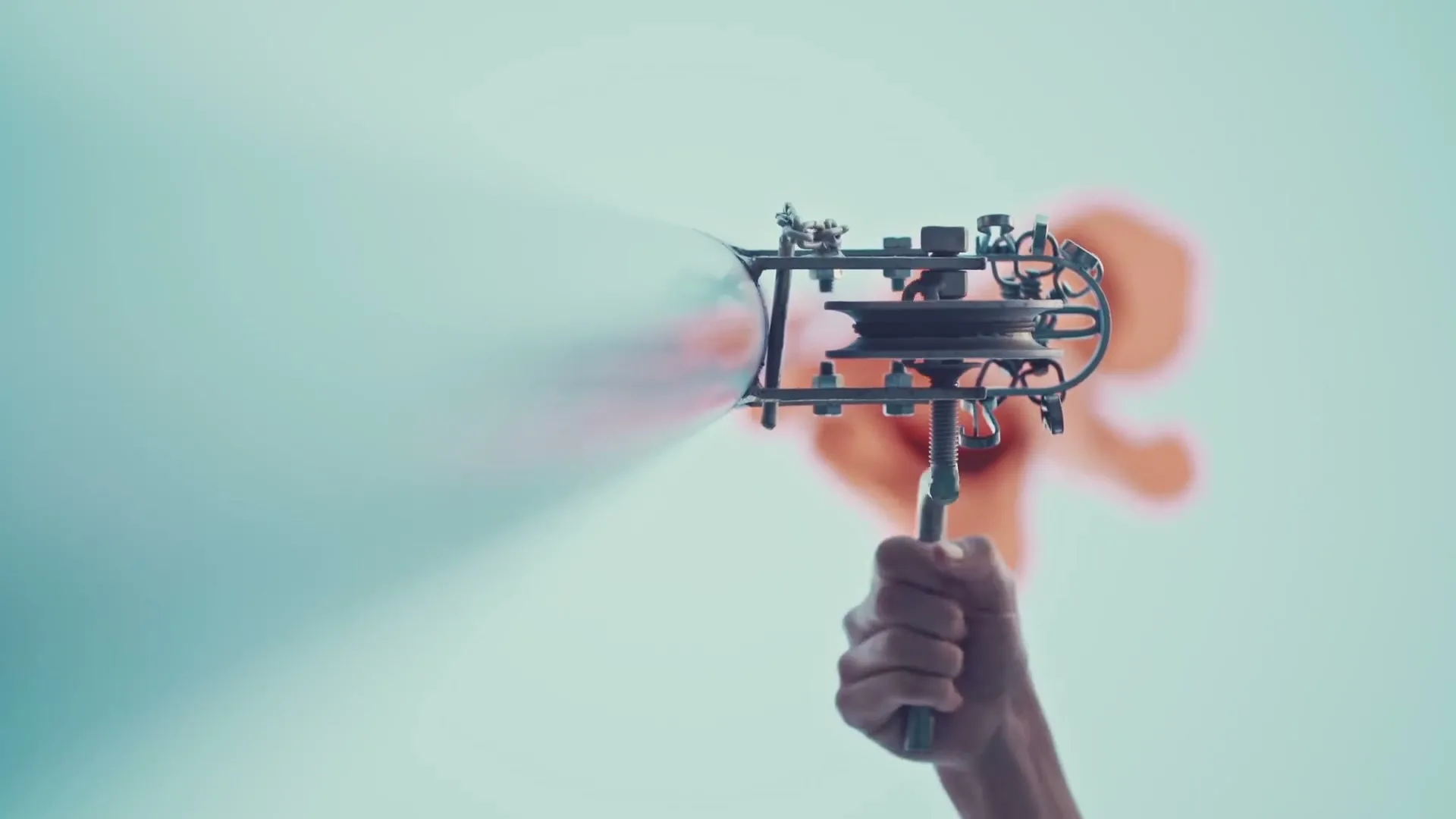 A person's hand holds a metallic, intricate device that projects a beam of light, with a blurred orange shape in the background.
