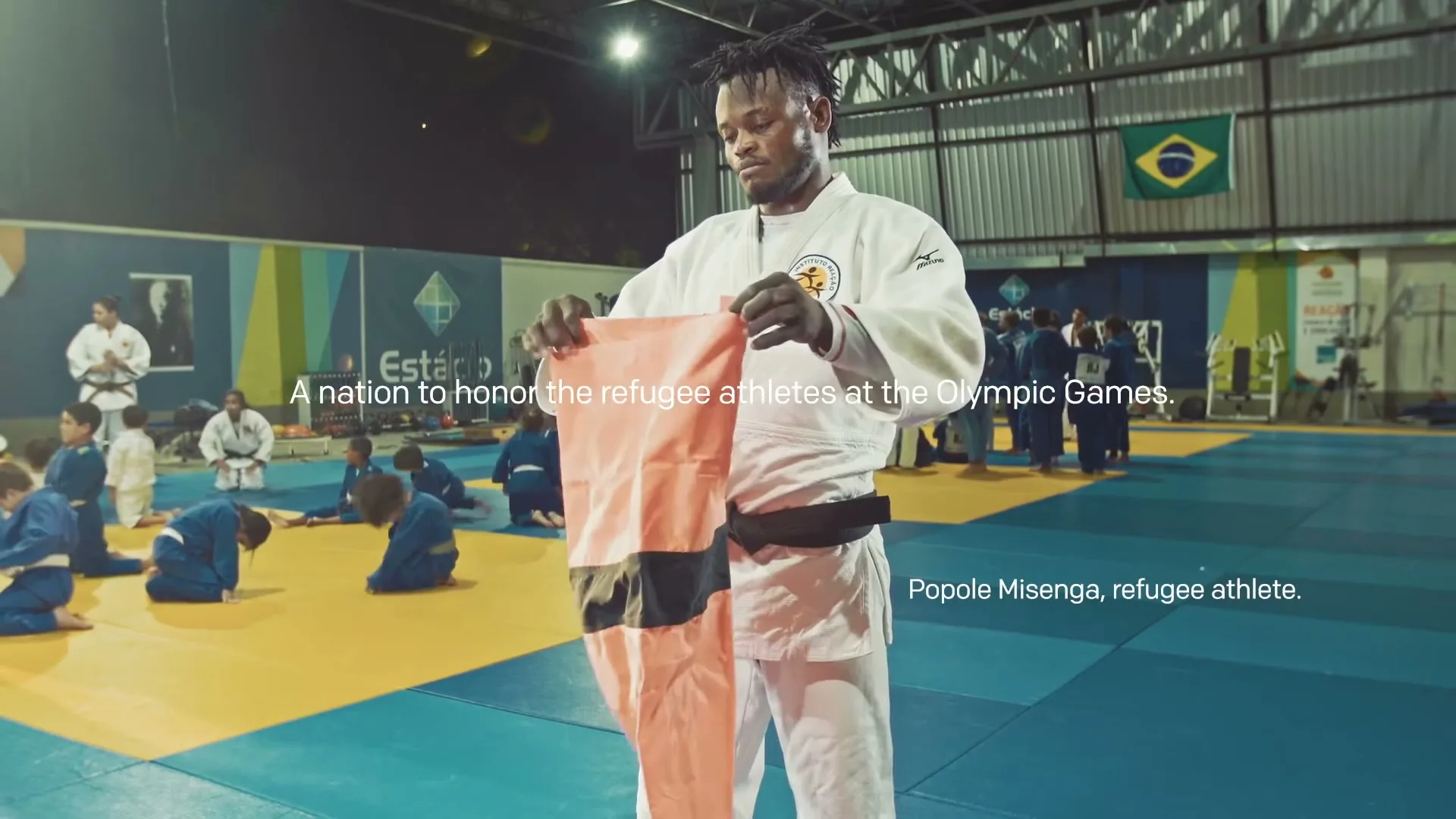 A male refugee athlete in a white gi holds up a flag with a black stripe, while children in blue gis practice judo in a gym.