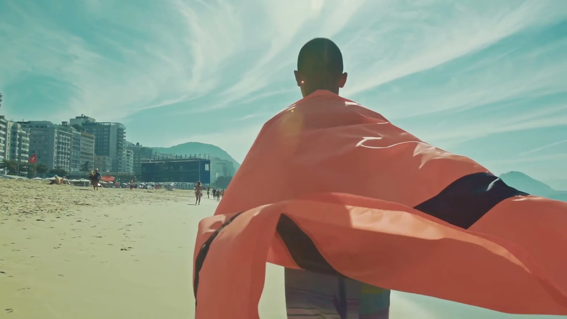 A young boy, seen from behind, runs along a sunny beach with a large orange and black flag draped over his shoulders, resembling a cape.