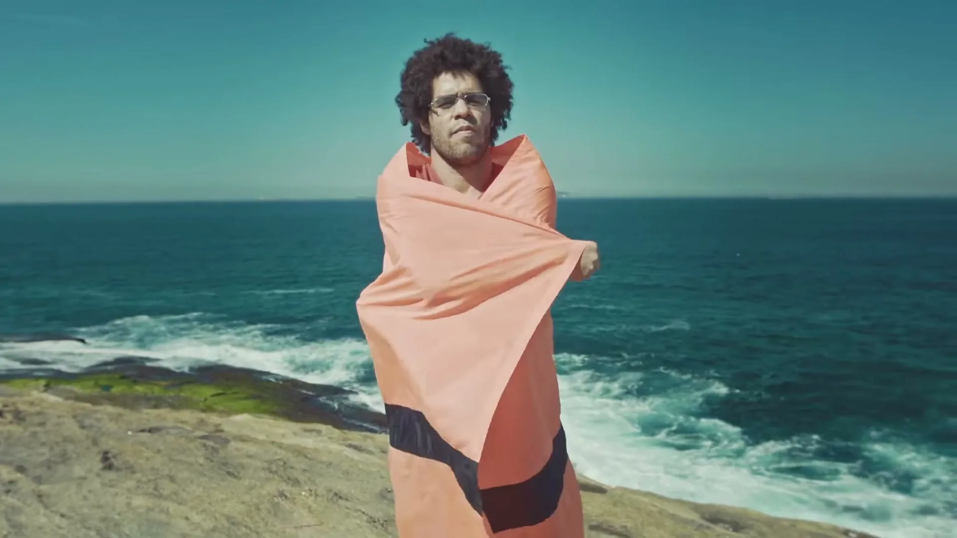 A man with curly hair and glasses, wrapped in an orange and black flag, stands on a rocky cliff overlooking the ocean on a sunny day.