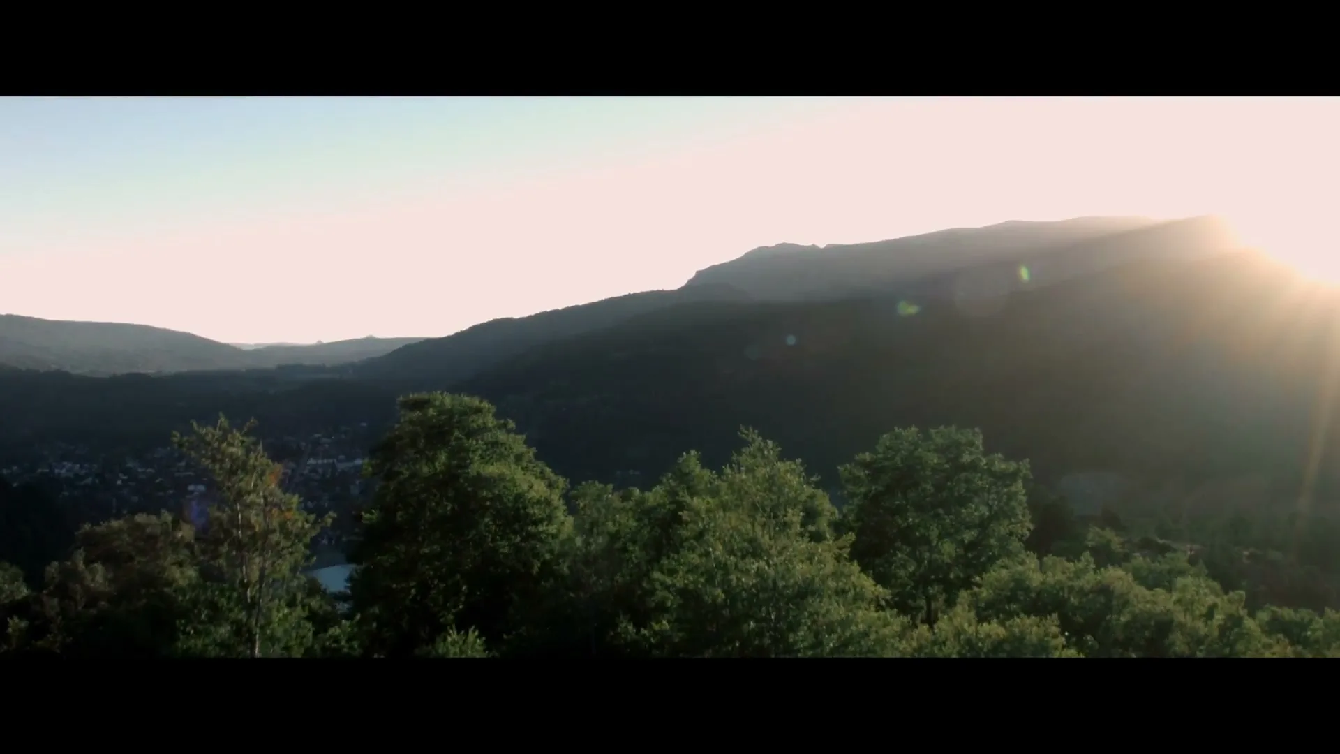 An aerial shot of a vast, green mountain landscape with a town and lake nestled in the valley, bathed in the warm glow of a setting sun.