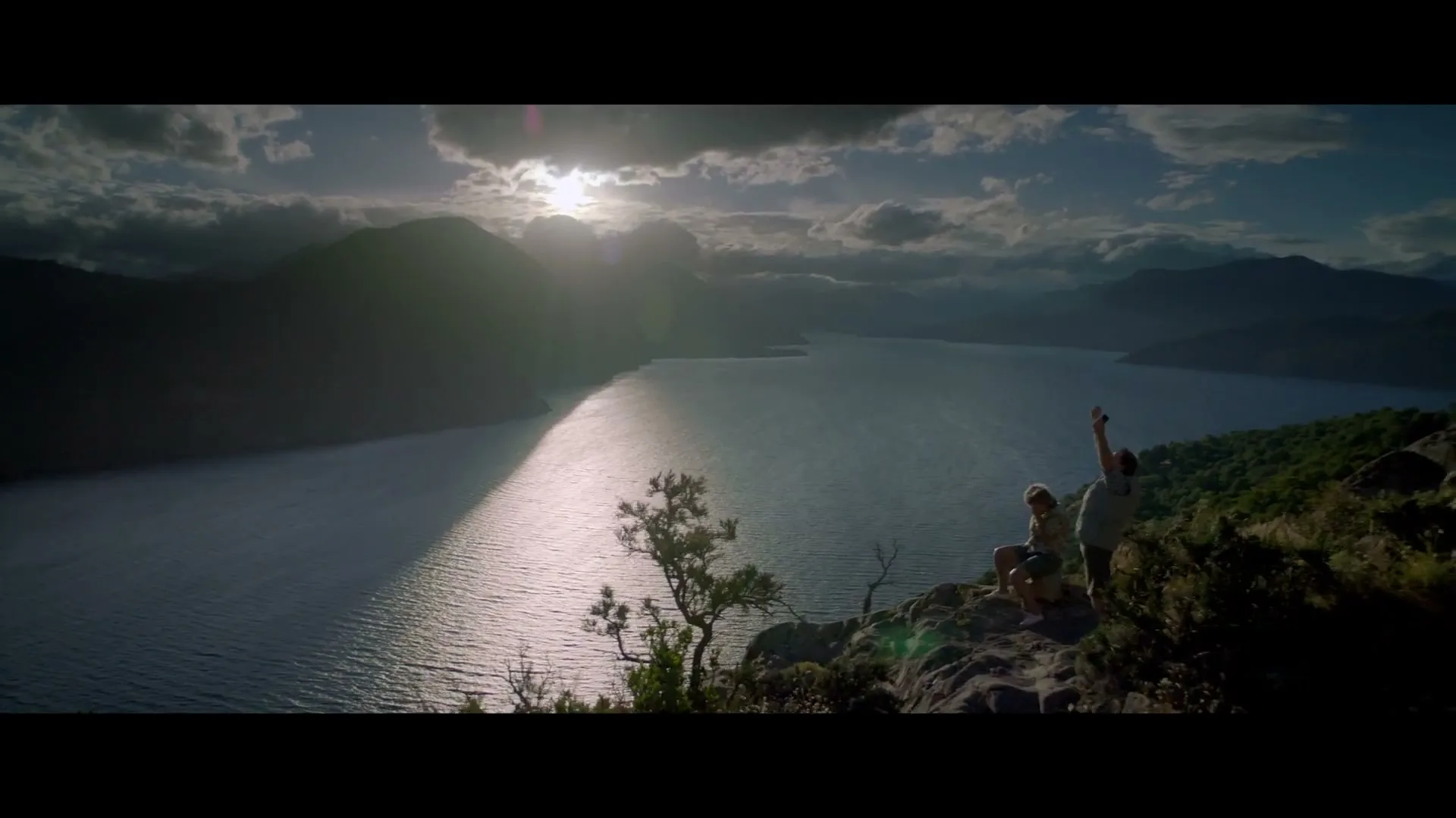 Two people sit on a rocky cliff overlooking a vast lake surrounded by mountains, with the sun setting over the horizon.