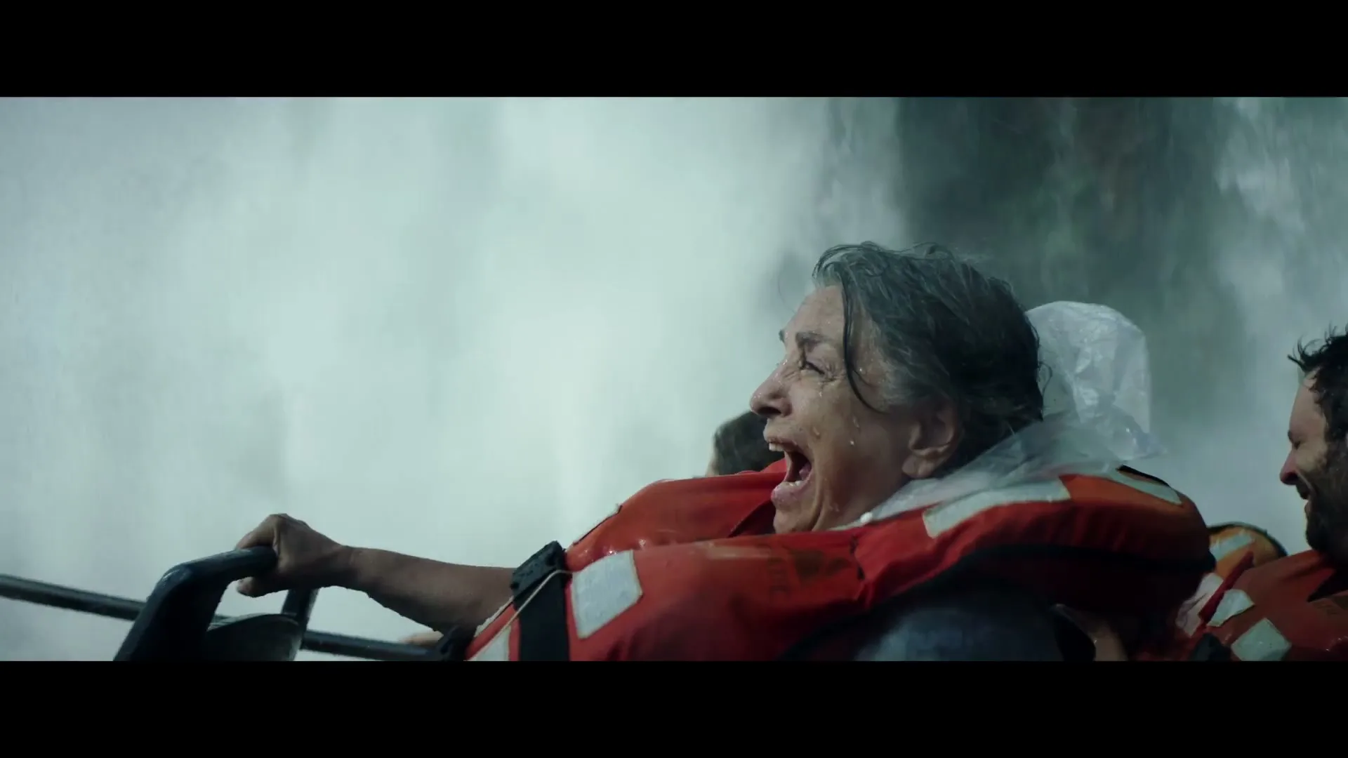 An older woman in a life vest screams with joy as she is drenched by a powerful waterfall, holding onto a boat railing.