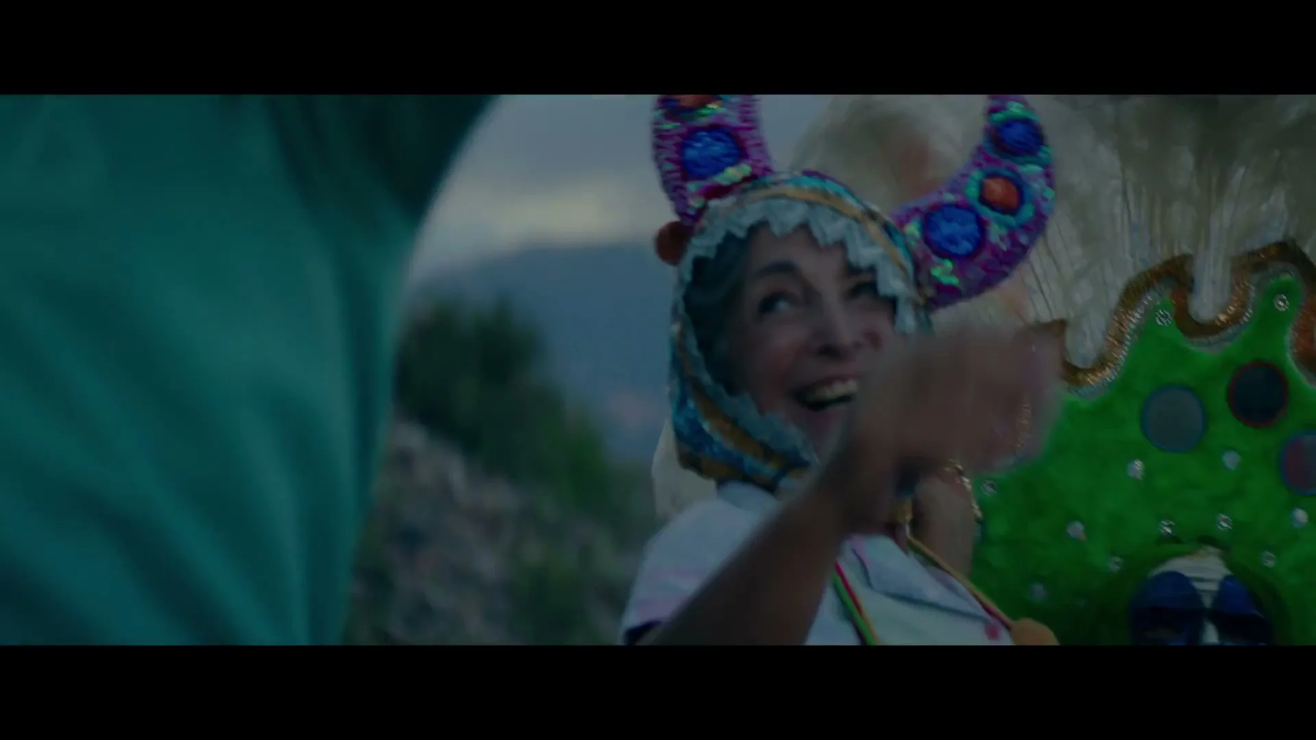 A joyful older woman in a colorful, horned headdress and white shirt laughs while looking up, with a blurred person in a teal shirt beside her and mountains in the background.