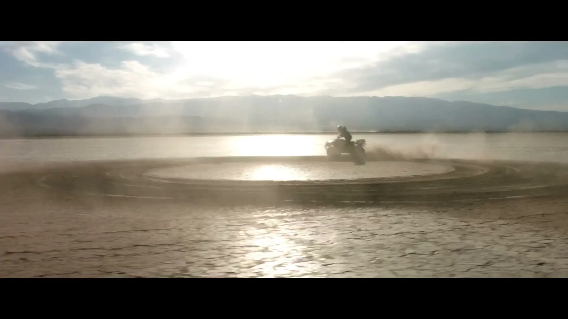 A person on a motorcycle rides in circles on a vast, cracked dry lakebed, kicking up dust under a bright sky.