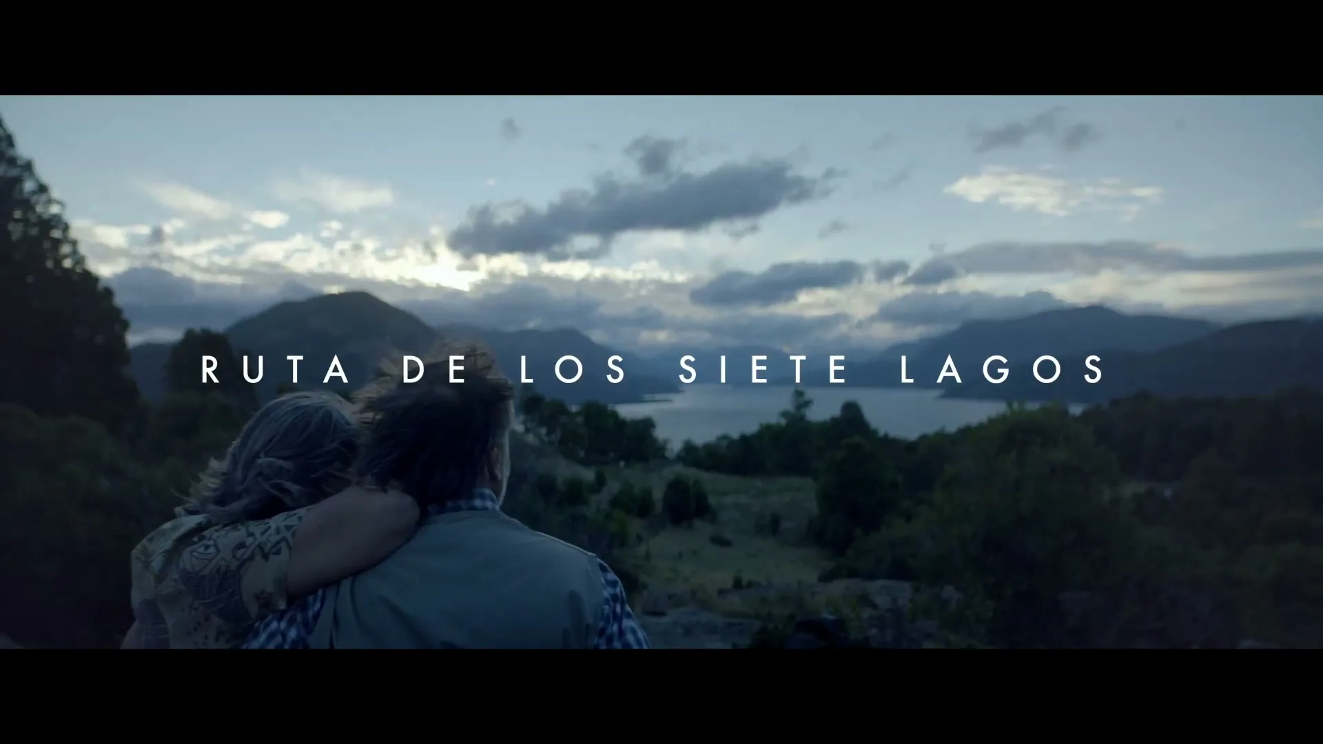 An older couple, a woman with grey hair in a floral top and a man in a plaid shirt and vest, embrace and look out at a lake and mountains at dusk.