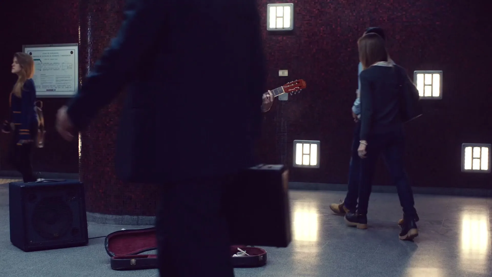 A bearded man plays an acoustic guitar in a subway station, with an open guitar case on the ground collecting money, as people walk past him.