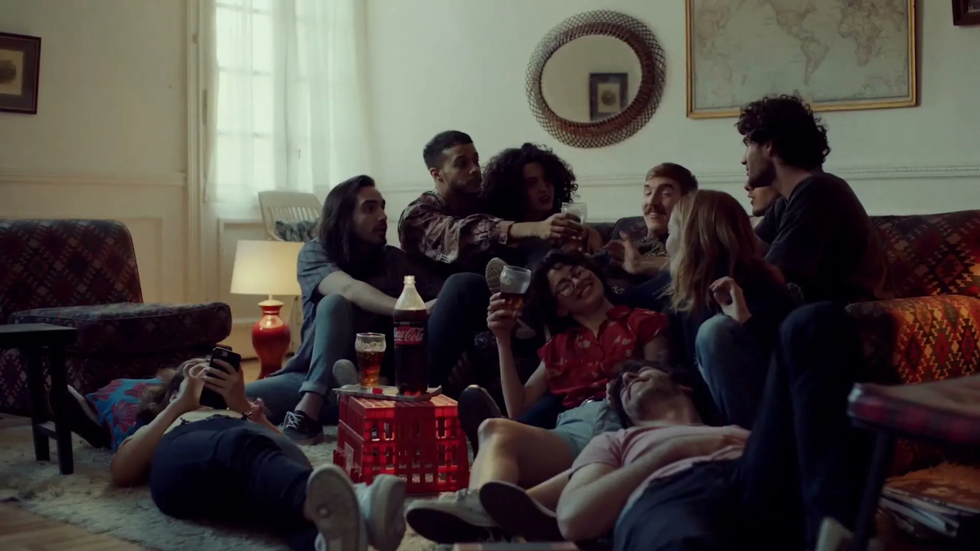 A diverse group of young adults relax and socialize in a cozy living room, some on the couch, others on the floor, with Coca-Cola bottles nearby.