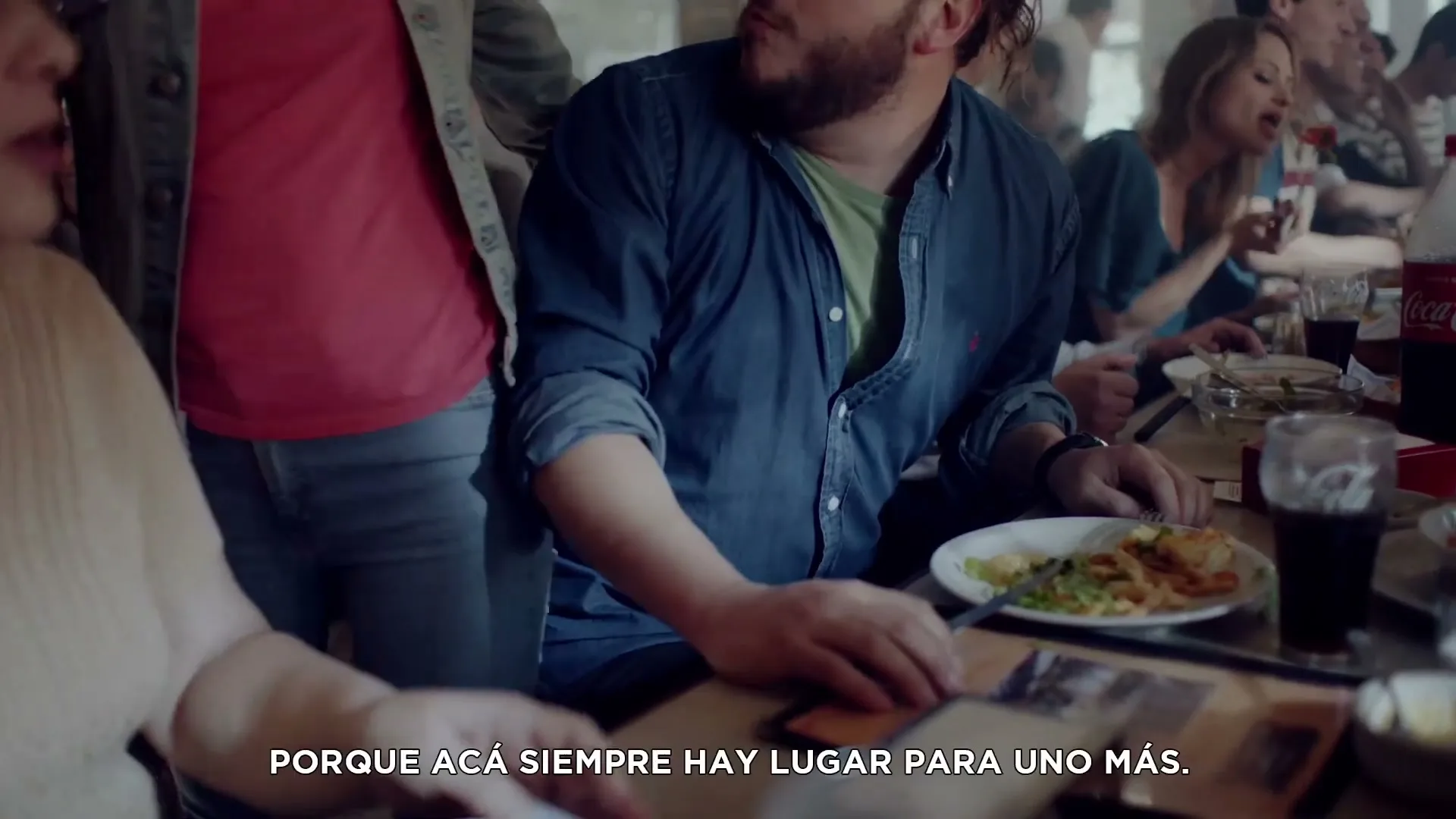 A diverse group of people are eating and talking at a crowded restaurant table, with a man in a blue shirt taking a large bite of food.