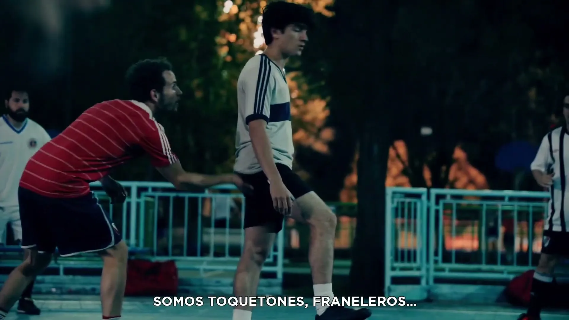 A group of men play soccer on an outdoor court at night, with one man in a red and white striped shirt reaching out to another.