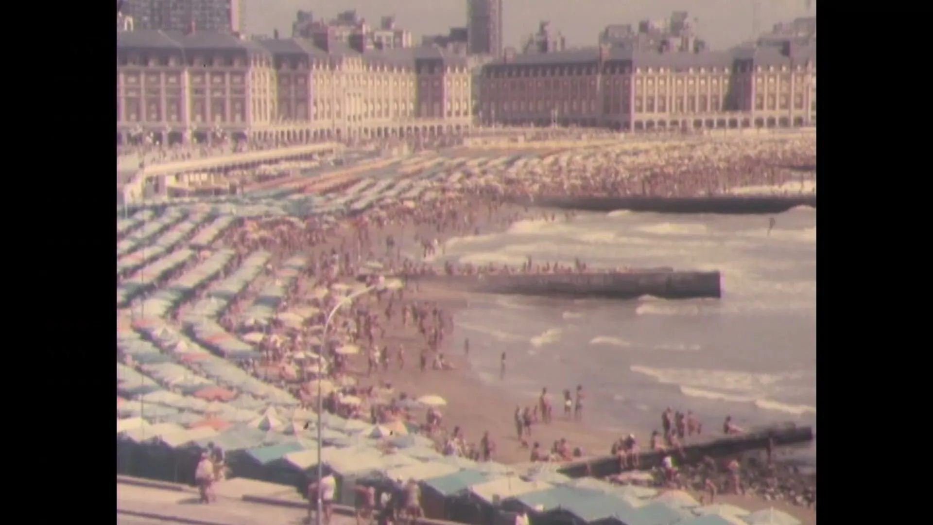 A wide shot captures a bustling beach scene with numerous people enjoying the sun and waves, while large buildings line the distant horizon.