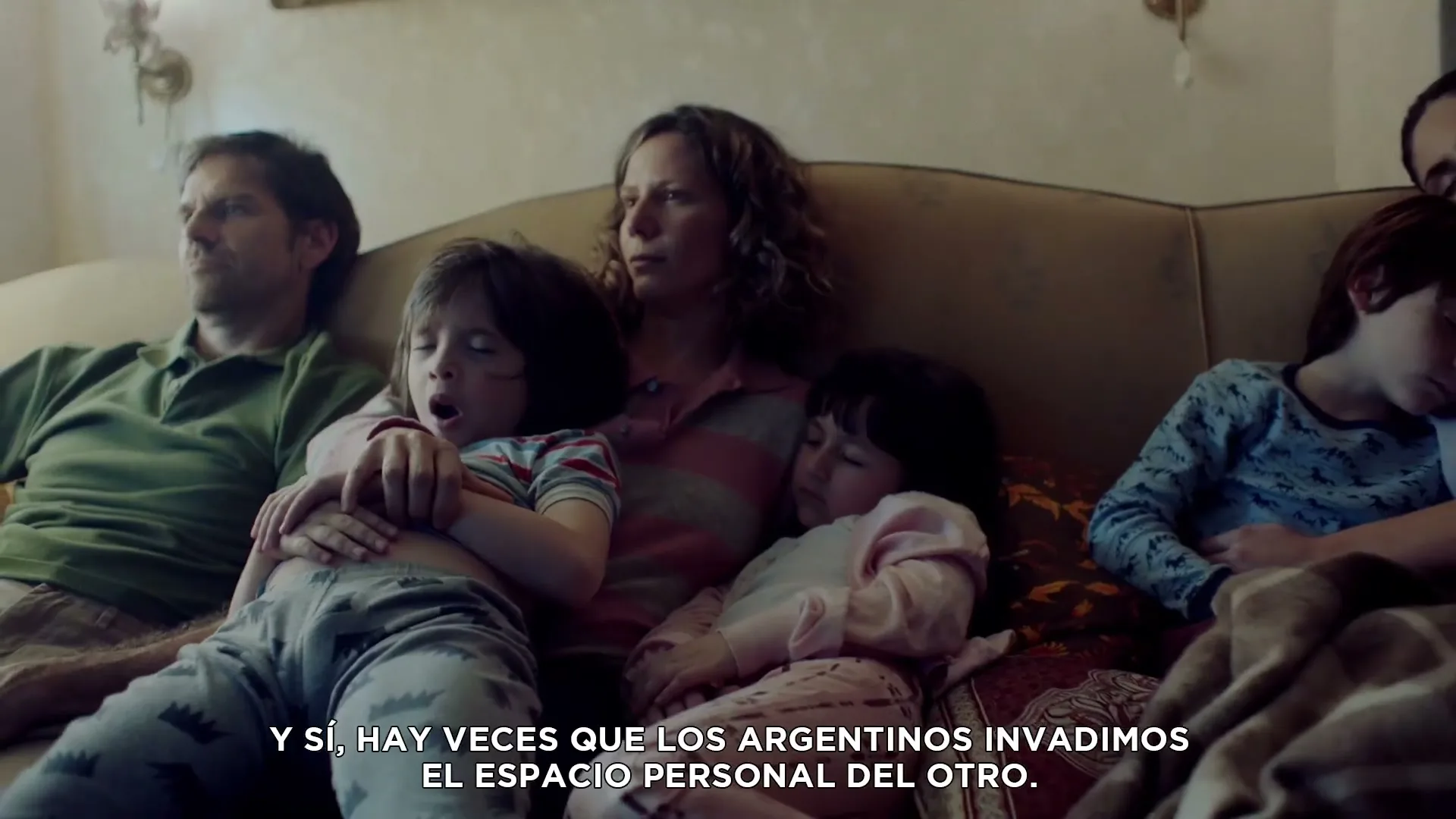 A family of five, including two adults and three children, are seen sleeping or resting on a couch in a cozy living room, illuminated by natural light.