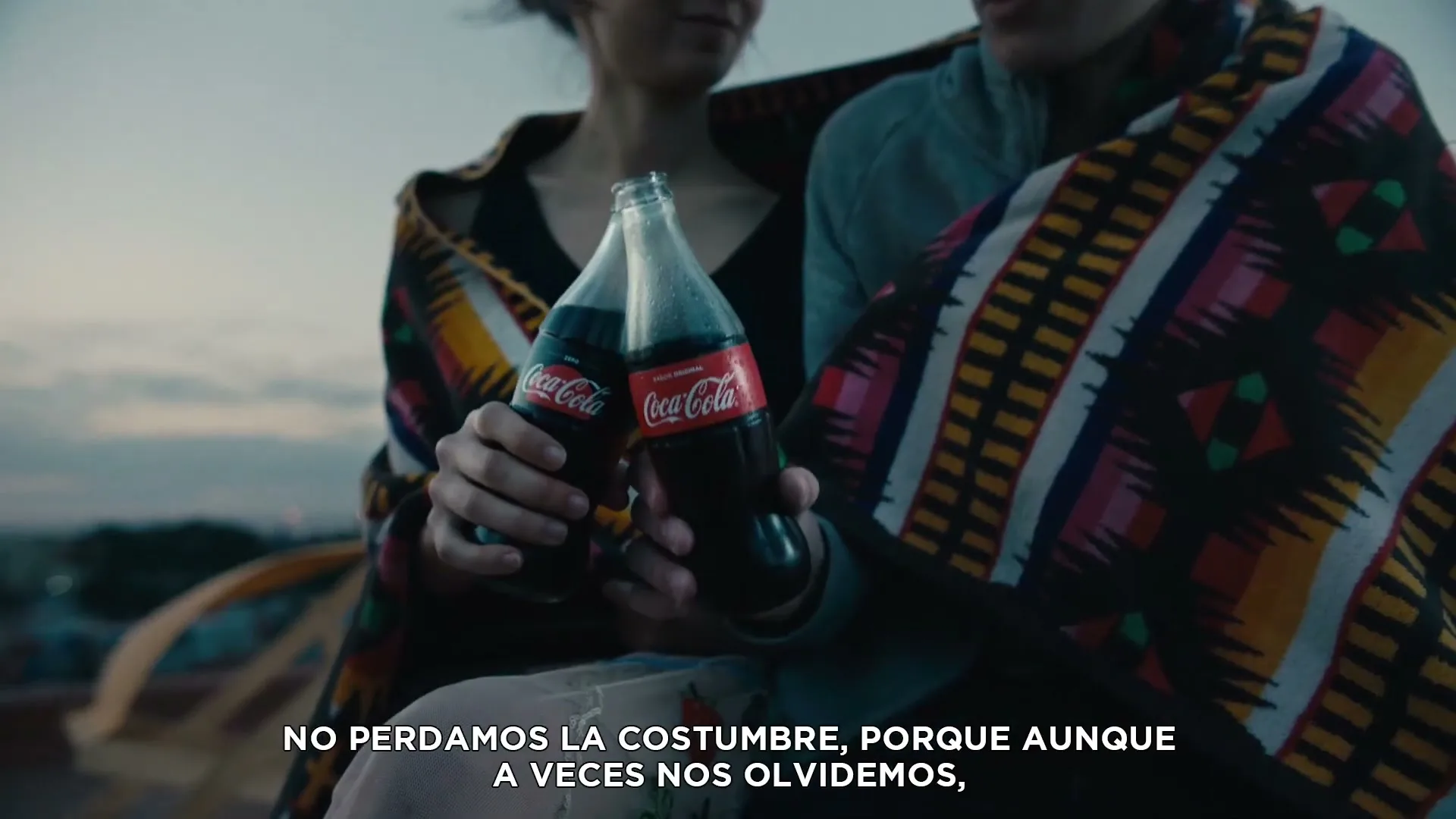 Two young adults, wrapped in a colorful blanket, hold up two Coca-Cola bottles and clink them together on a rooftop at dusk.