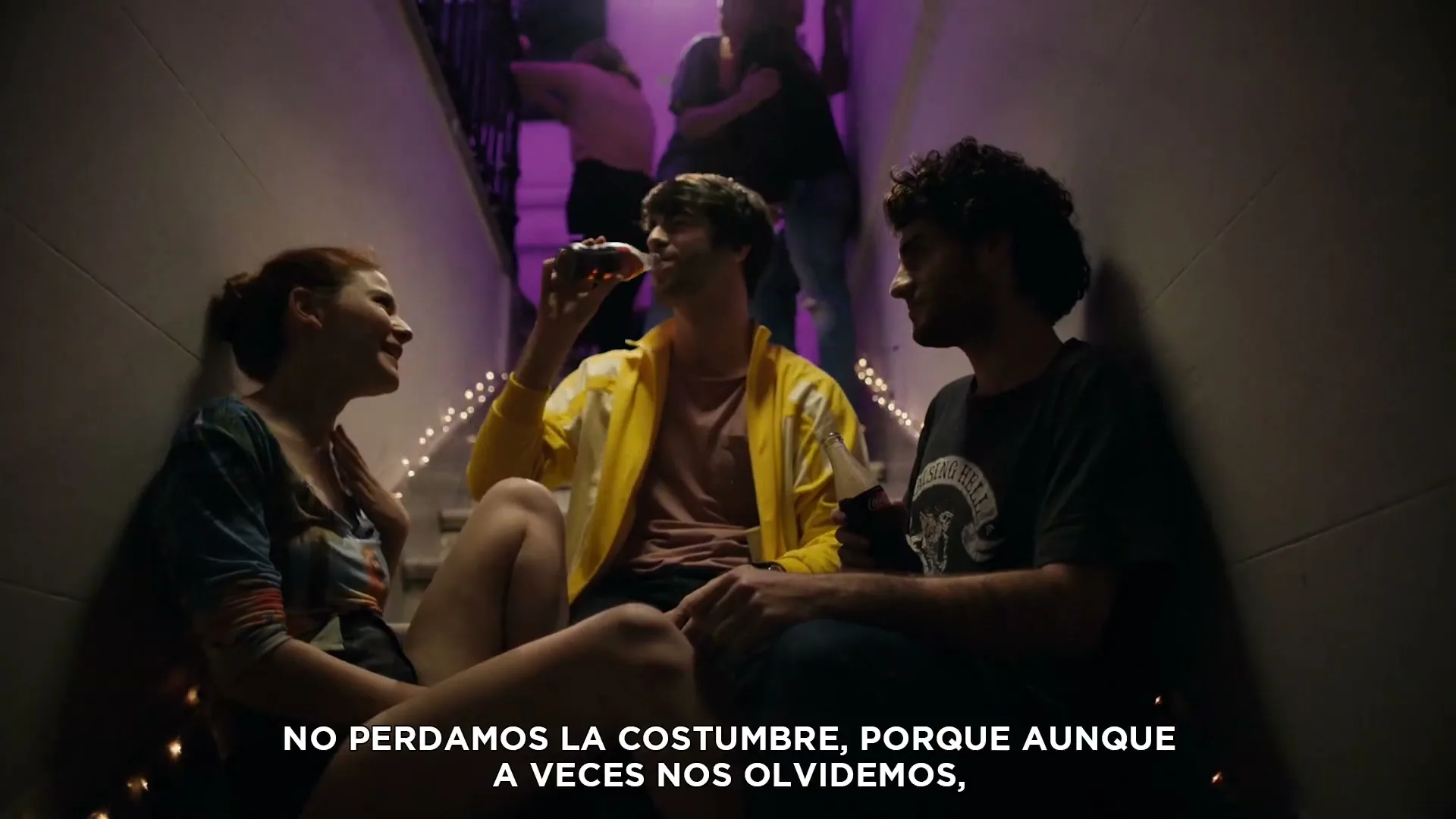 Three young adults sit on a staircase, talking and drinking Coca-Cola, while other people socialize in the background at a party.