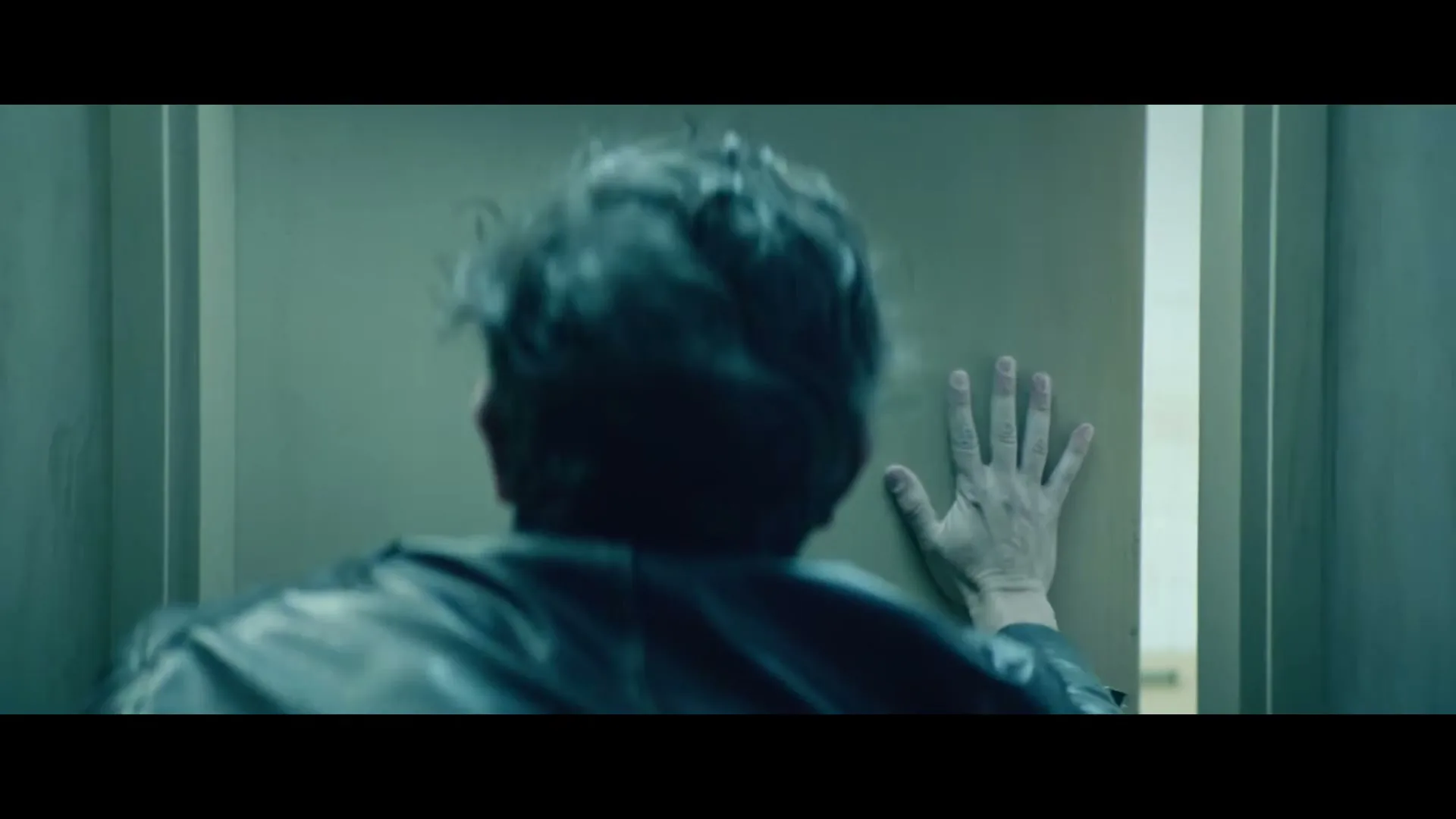 A man with dark, messy hair and a black jacket stumbles towards a men's restroom door, his hand outstretched, in a dimly lit hallway.