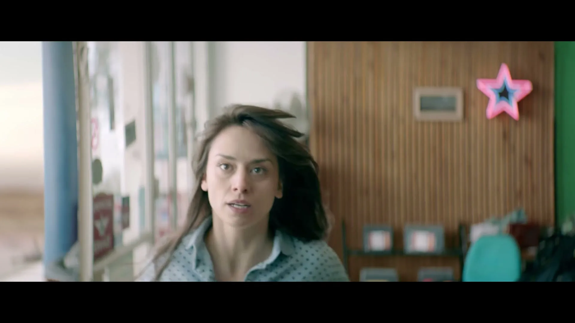 A woman with long brown hair and a polka dot shirt runs towards the camera in a brightly lit room with a wooden wall and a pink star decoration.