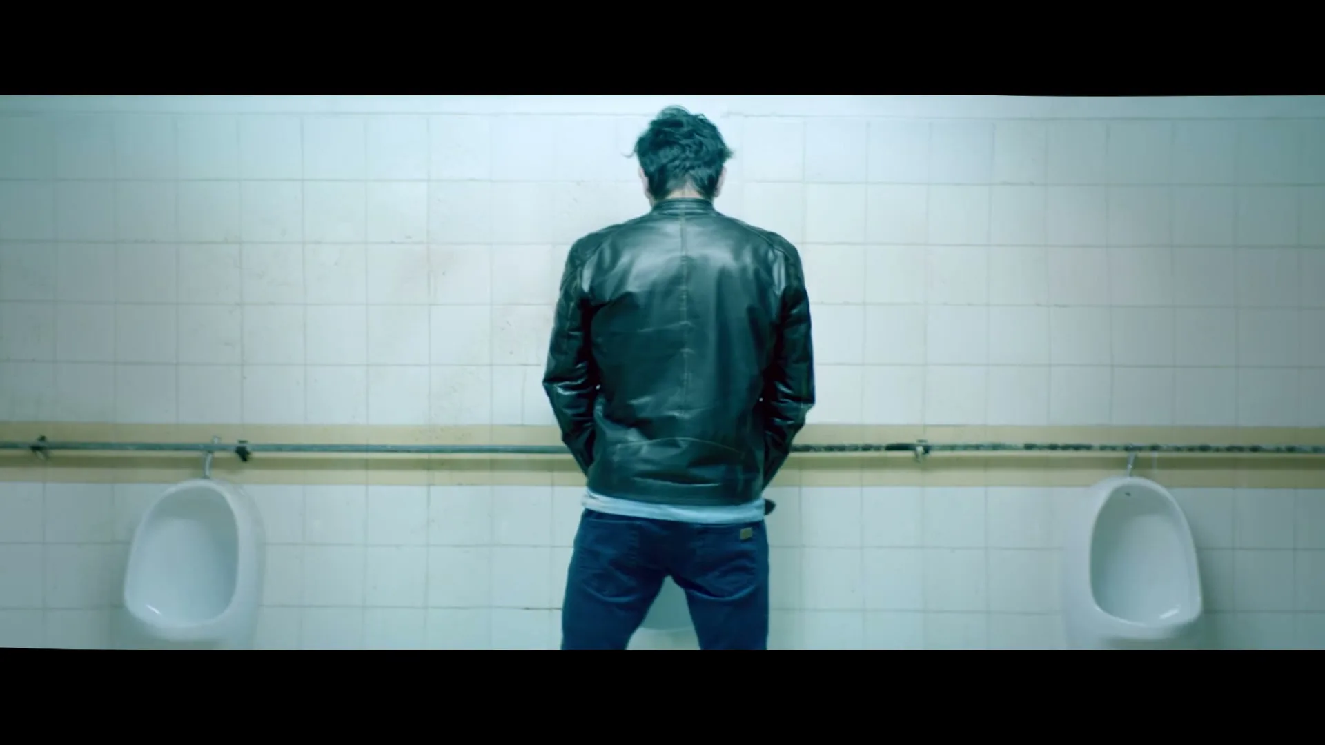 A man in a black leather jacket and jeans stands with his back to the camera, urinating in a public restroom.