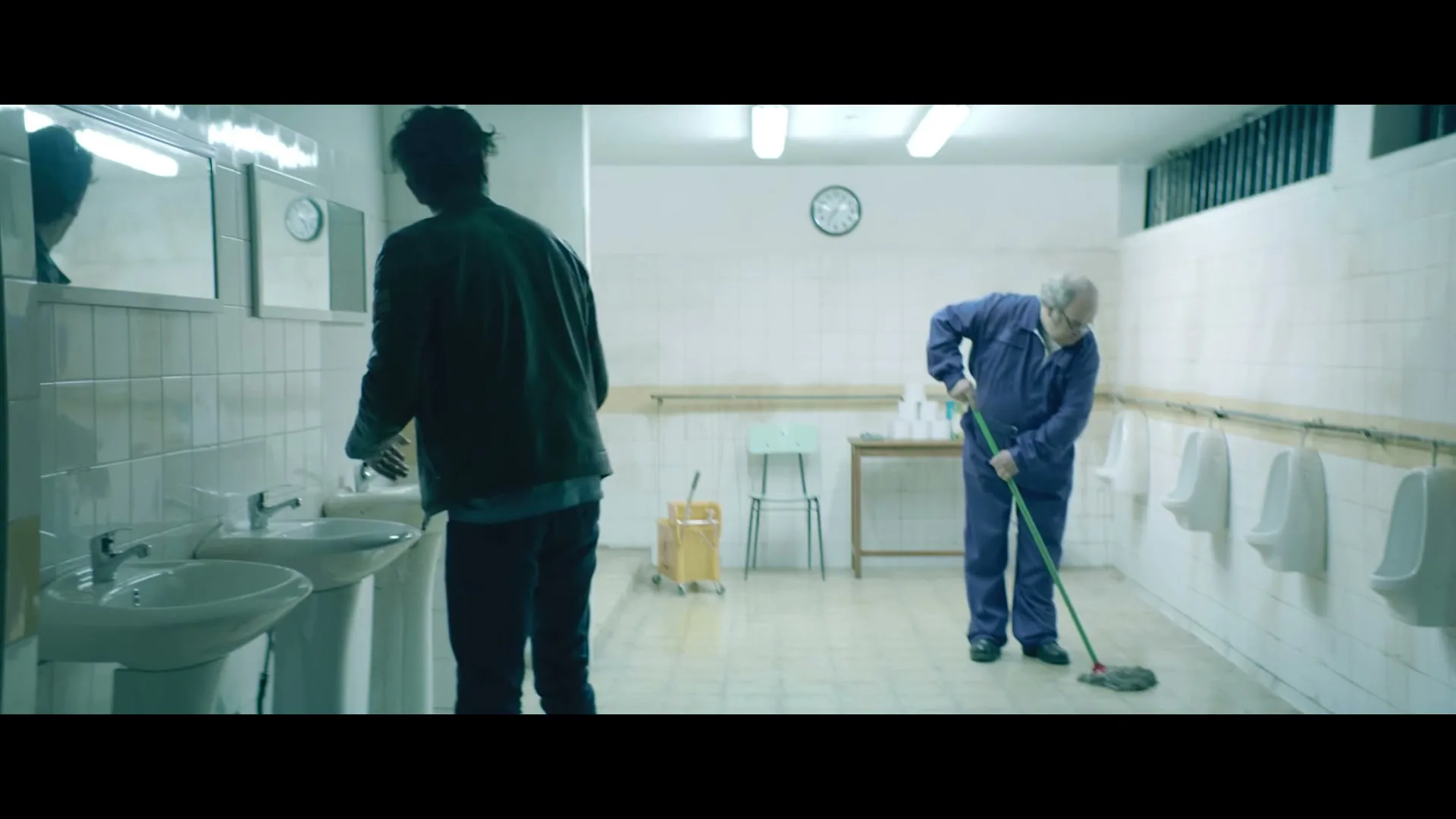 A man washes his hands at a sink while an older man mops the floor in a public restroom with urinals.