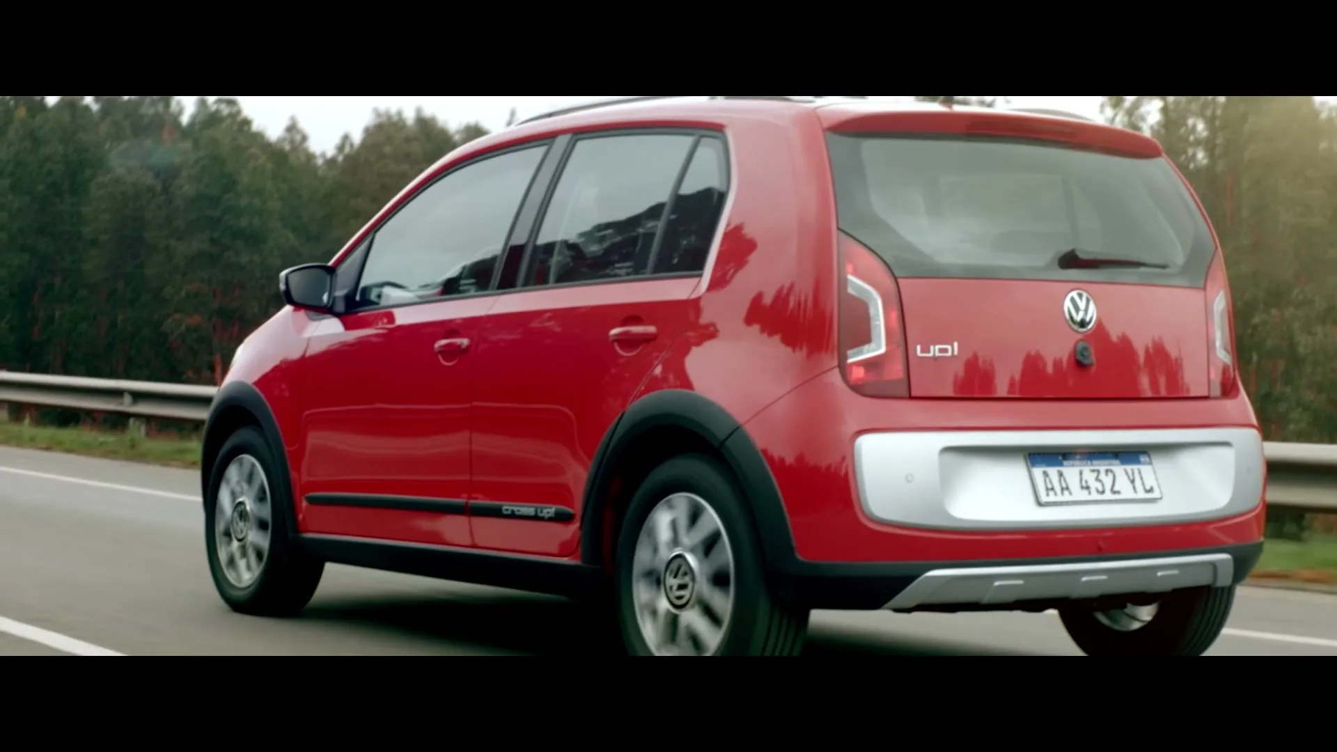 A red Volkswagen Cross Up! drives away from the camera on a highway, with trees in the background under a bright sky.