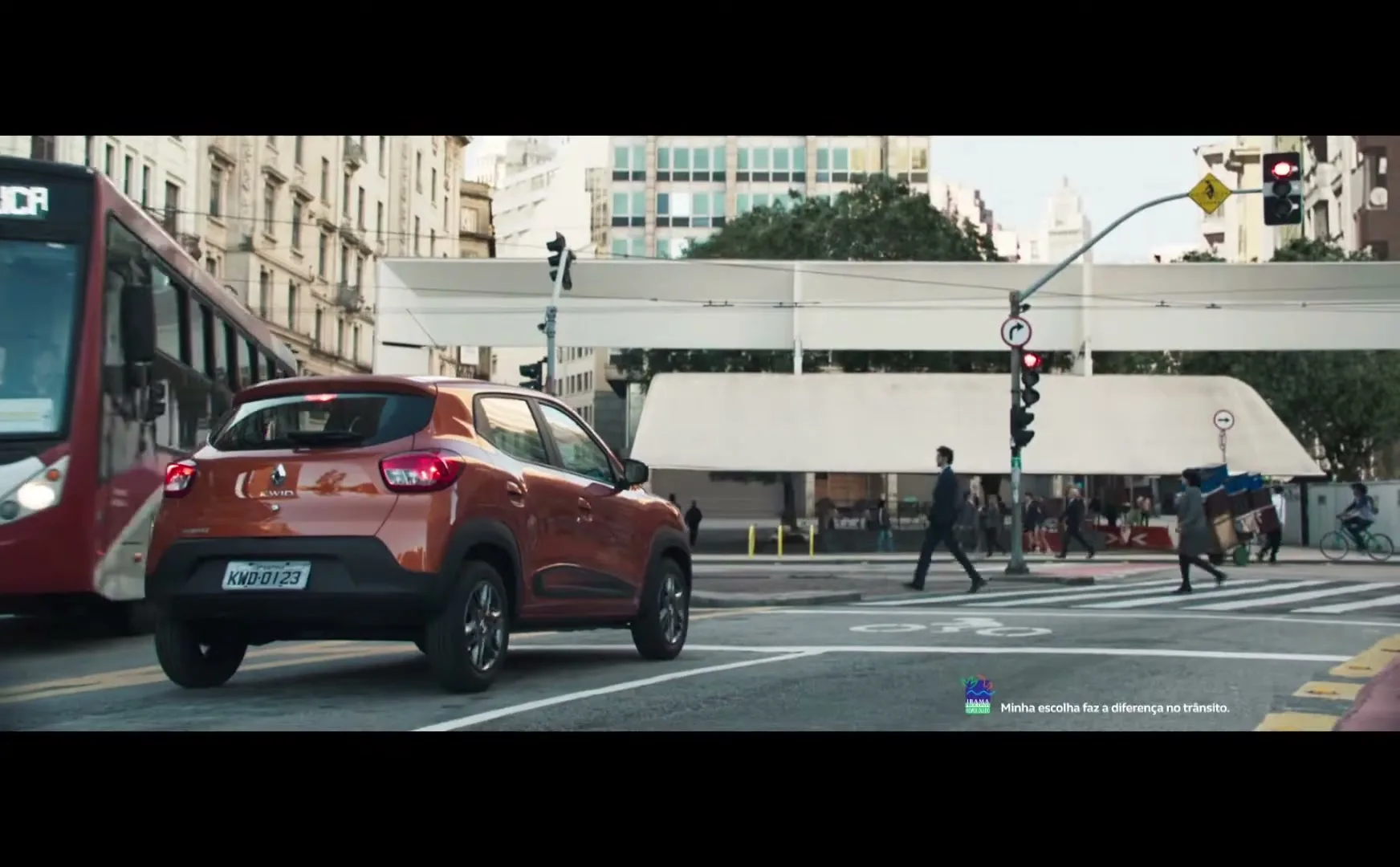 An orange Renault Kwid is stopped at a red light in a busy city intersection, with pedestrians crossing and a bus on the left.