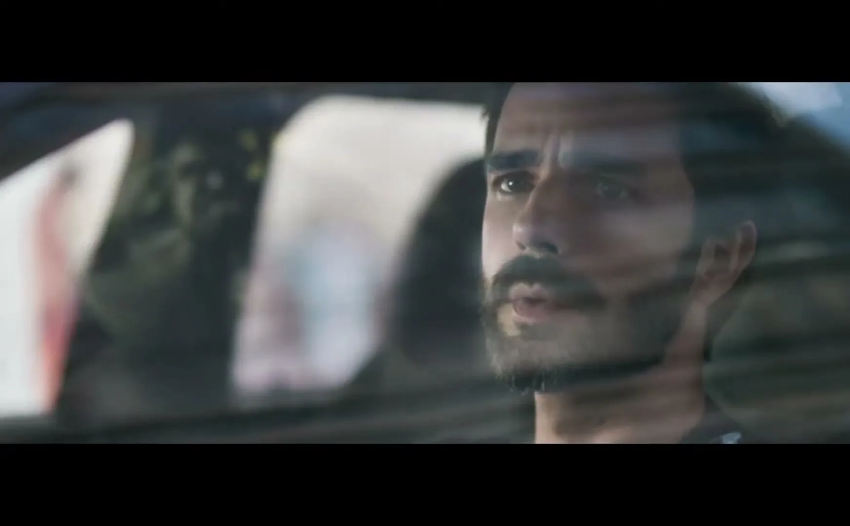A man with a beard and mustache looks out the window of a car, with reflections visible on the glass, in a medium closeup shot.