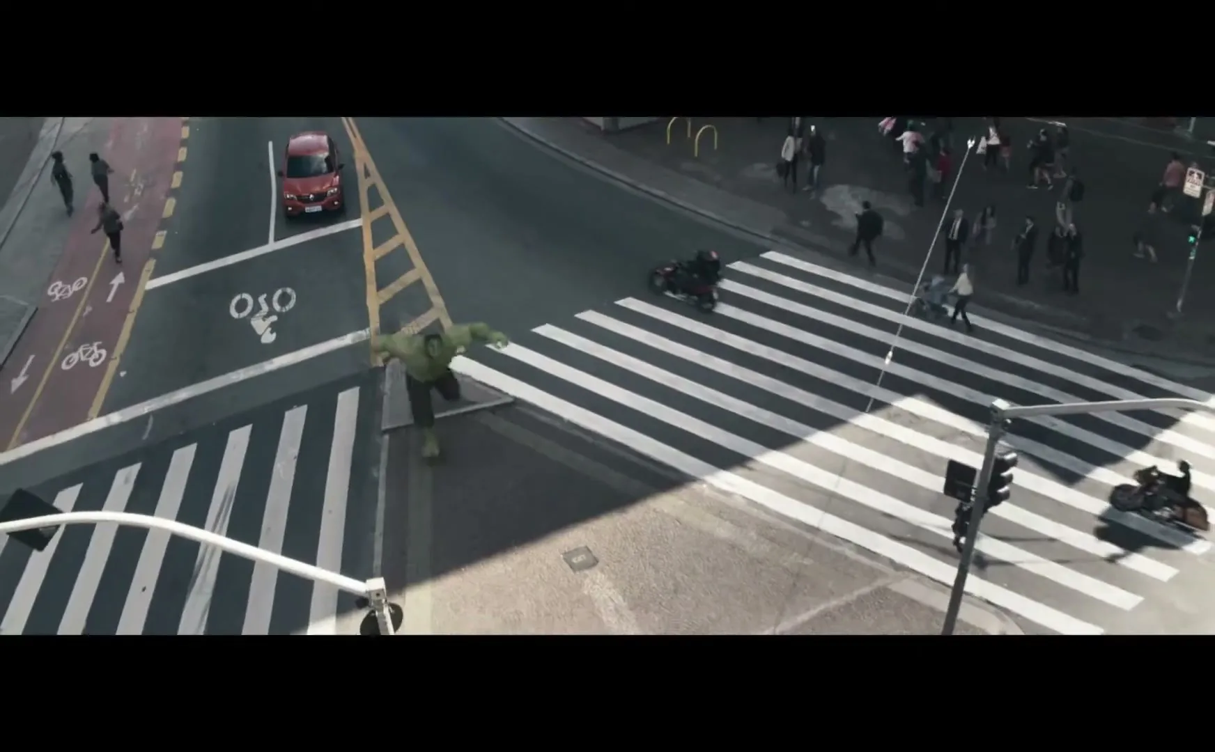 The Hulk, a large green CGI character, runs across a crosswalk in a busy city street, with a red car stopped nearby.
