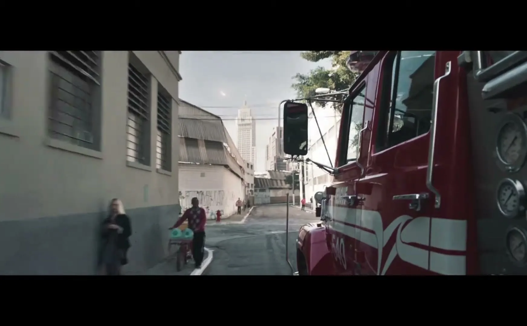 A red fire truck drives down a narrow street in a city, passing pedestrians and buildings under a cloudy sky.