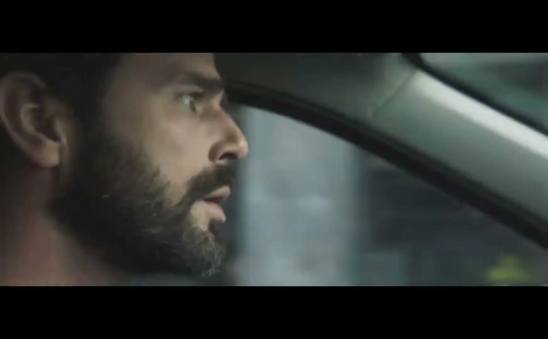 A man with a beard drives a car, looking surprised as he turns the steering wheel, in a medium closeup shot.