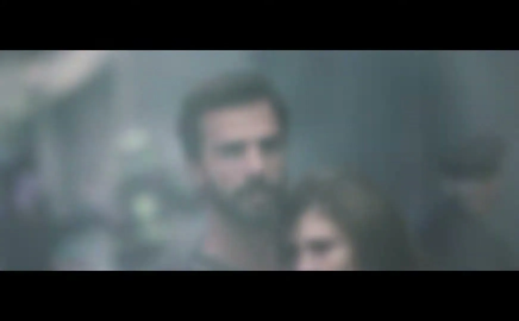 A man with a beard and a woman with long hair embrace in a smoky, dimly lit interior, looking serious.