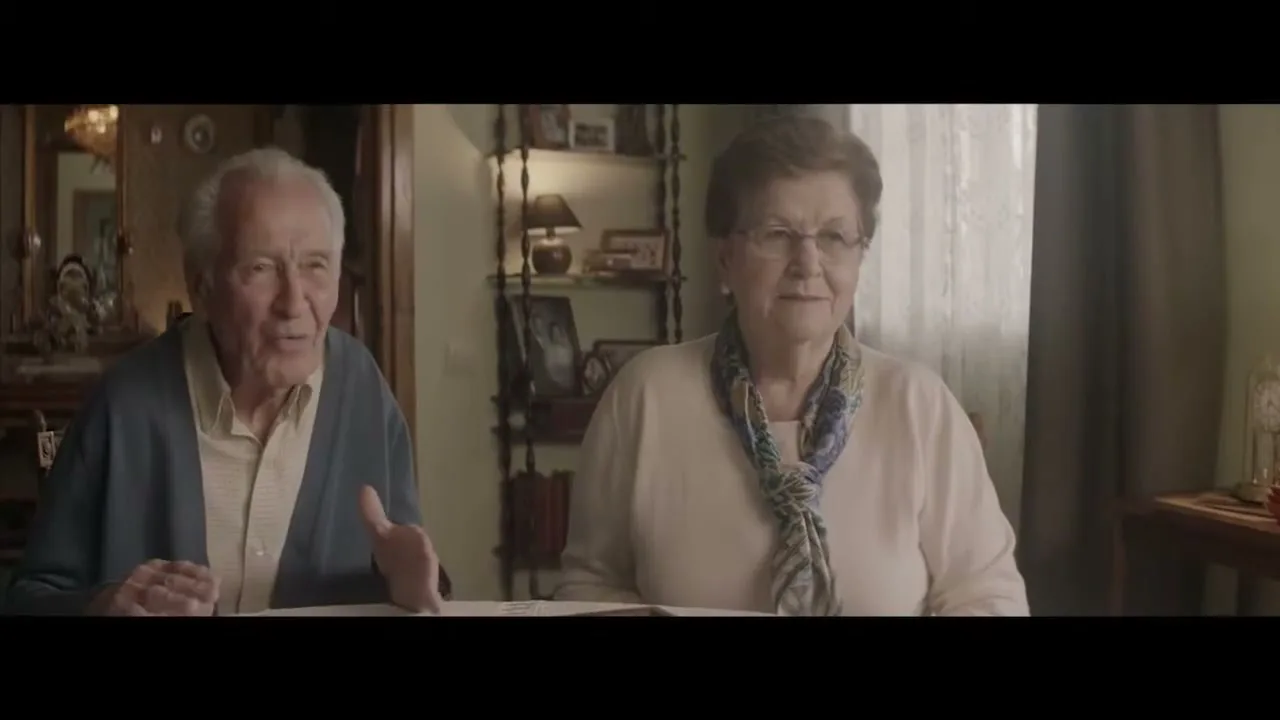 An elderly man and woman sit at a table, looking forward and talking, with the title "EL BESO" superimposed on screen.