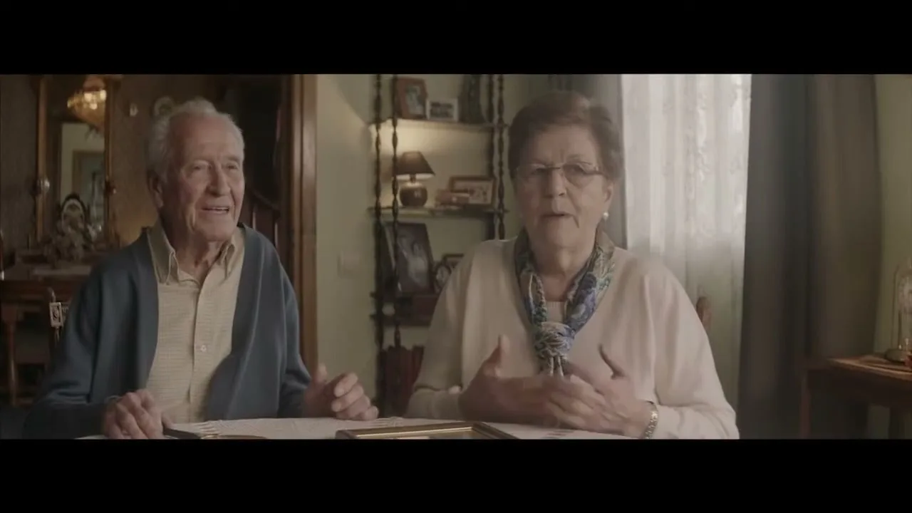 An elderly couple sits at a table in their home, the woman speaking and gesturing while the man listens and smiles.