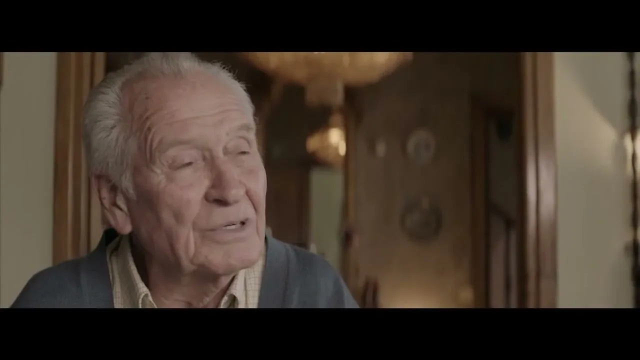 An elderly man with white hair and a blue cardigan speaks directly to the camera, smiling slightly in a warm, inviting interior setting.