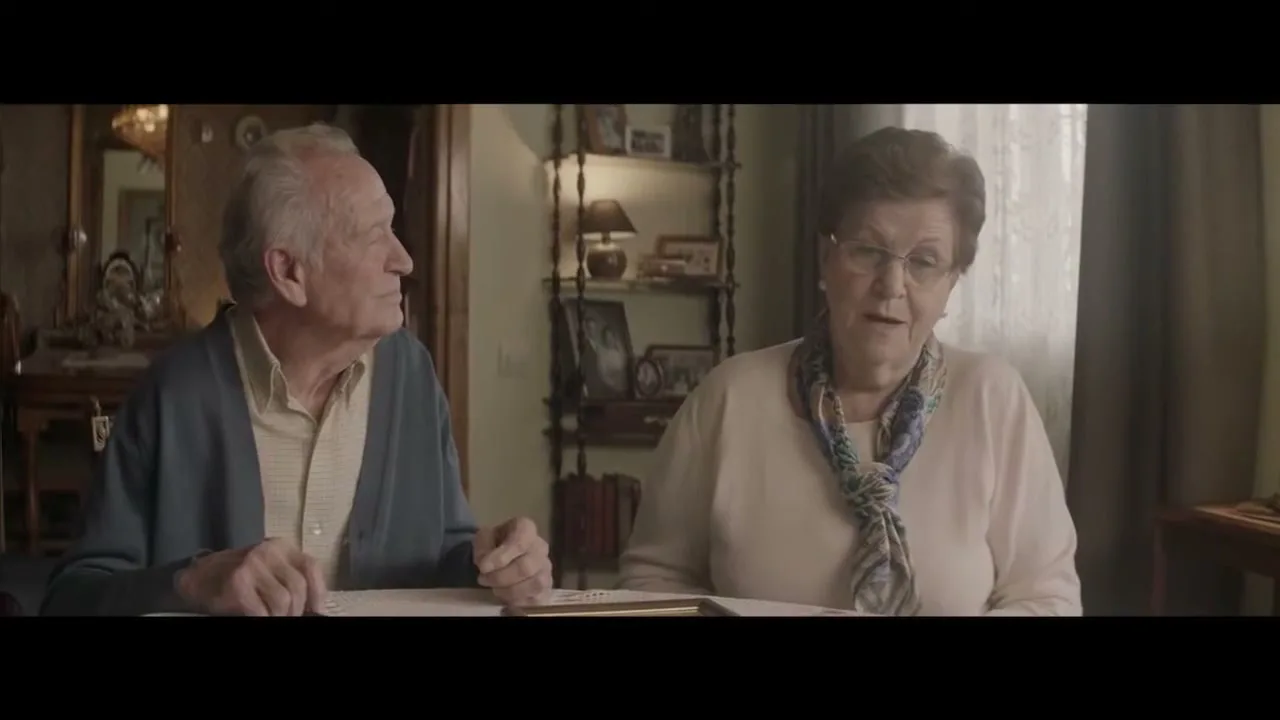 An elderly couple sits at a table in their home, the woman speaking while the man listens attentively, both looking off-camera.