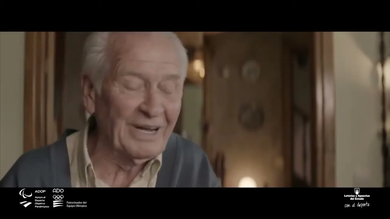 An elderly man with white hair and a blue cardigan speaks directly to the camera, smiling slightly in a warm, inviting interior setting.