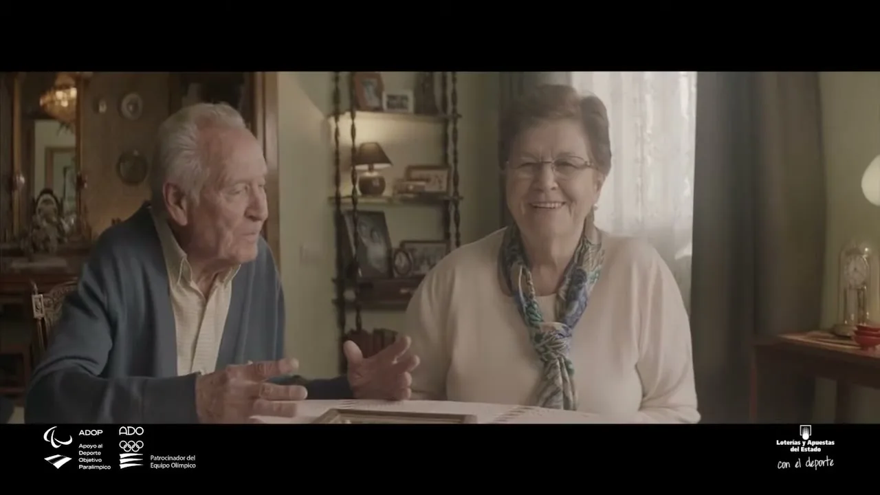 An elderly man in a blue cardigan and a woman in a cream top and scarf sit at a table, talking and laughing in a cozy living room.