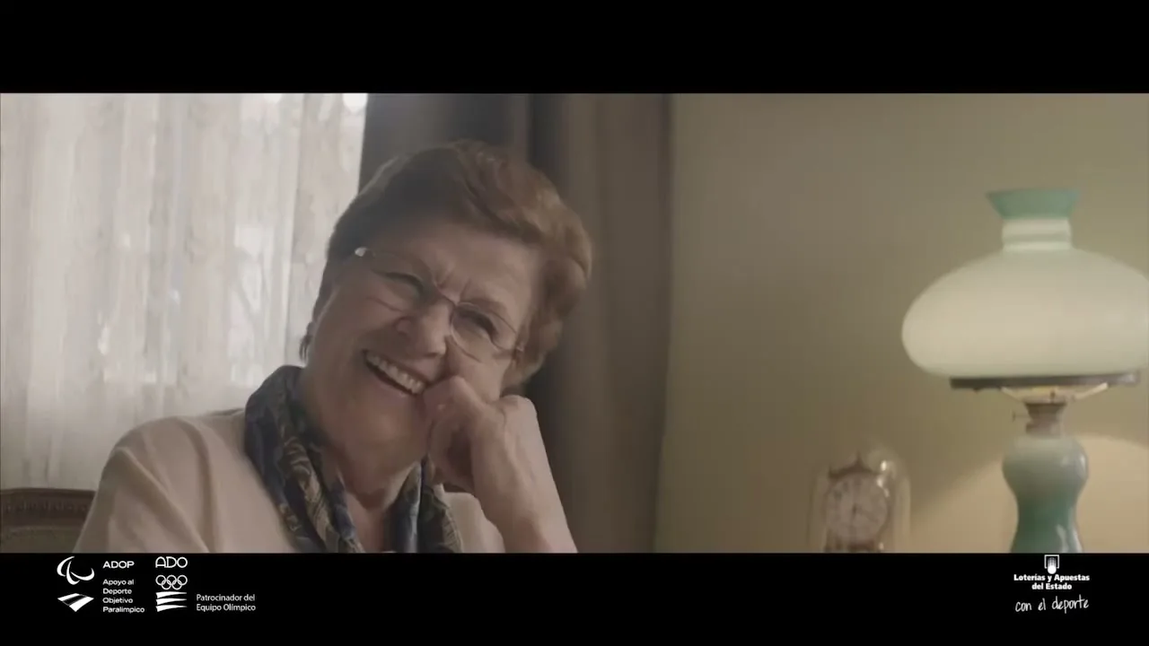 An elderly woman with short red hair and glasses, wearing a patterned scarf, smiles and laughs while sitting in a cozy living room.