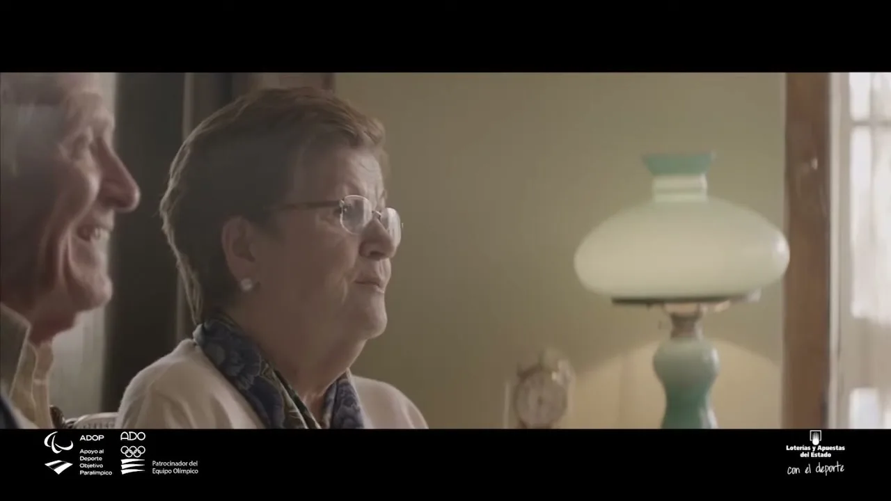 An elderly woman with glasses and a patterned scarf smiles while looking off-camera, with an elderly man partially visible beside her in a cozy room.